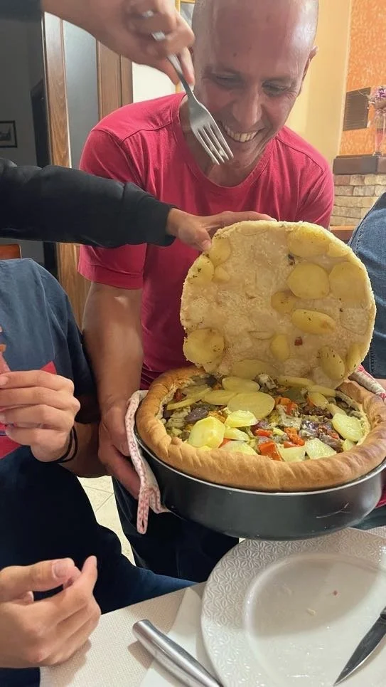 A man in a red shirt smiling as he lifts the lid off a large pizza, revealing toppings like potatoes, carrots, and other vegetables, at a restaurant.
