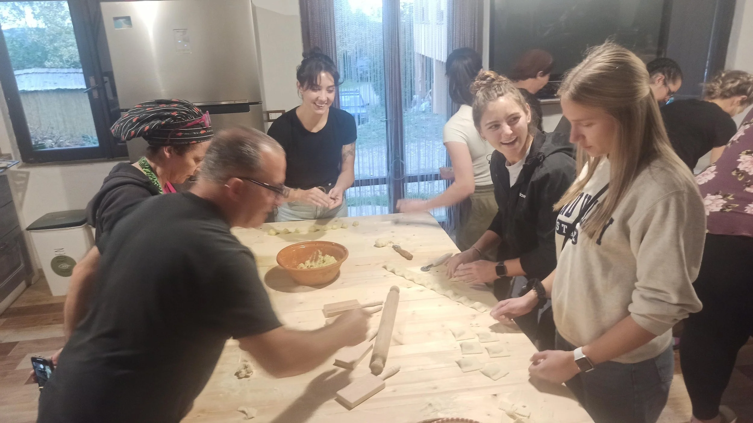 Group of people making dumplings together in a kitchen, with a woman in a headscarf in the foreground and others smiling and working on dough around a wooden table.
