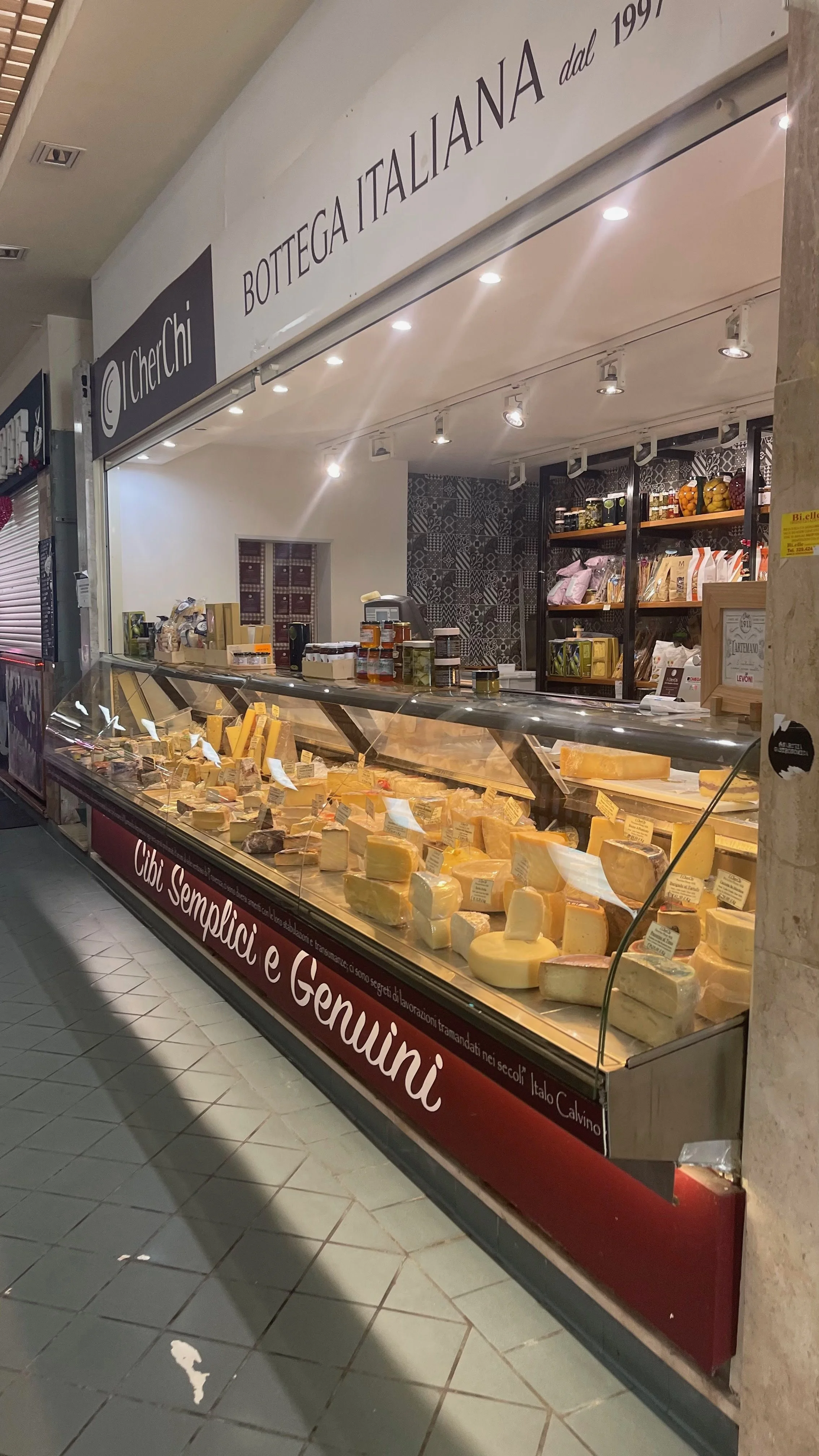 Display case with various cheeses at an Italian cheese shop, with shelves of jars and packaged goods behind the counter.