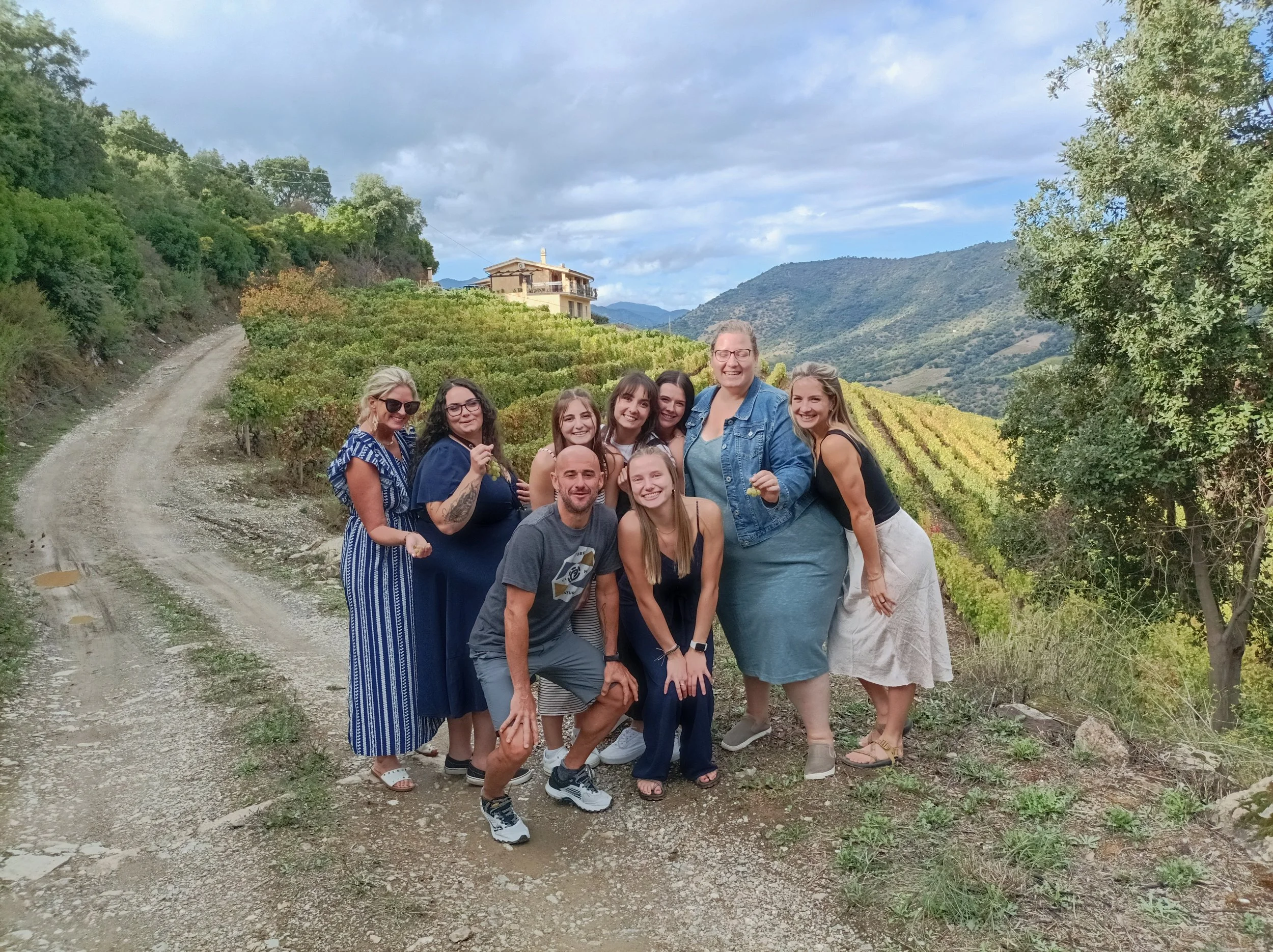 A group of nine people, including men and women, standing and kneeling together outdoors on a dirt path with terraced vineyards and rolling hills in the background. The sky is partly cloudy.