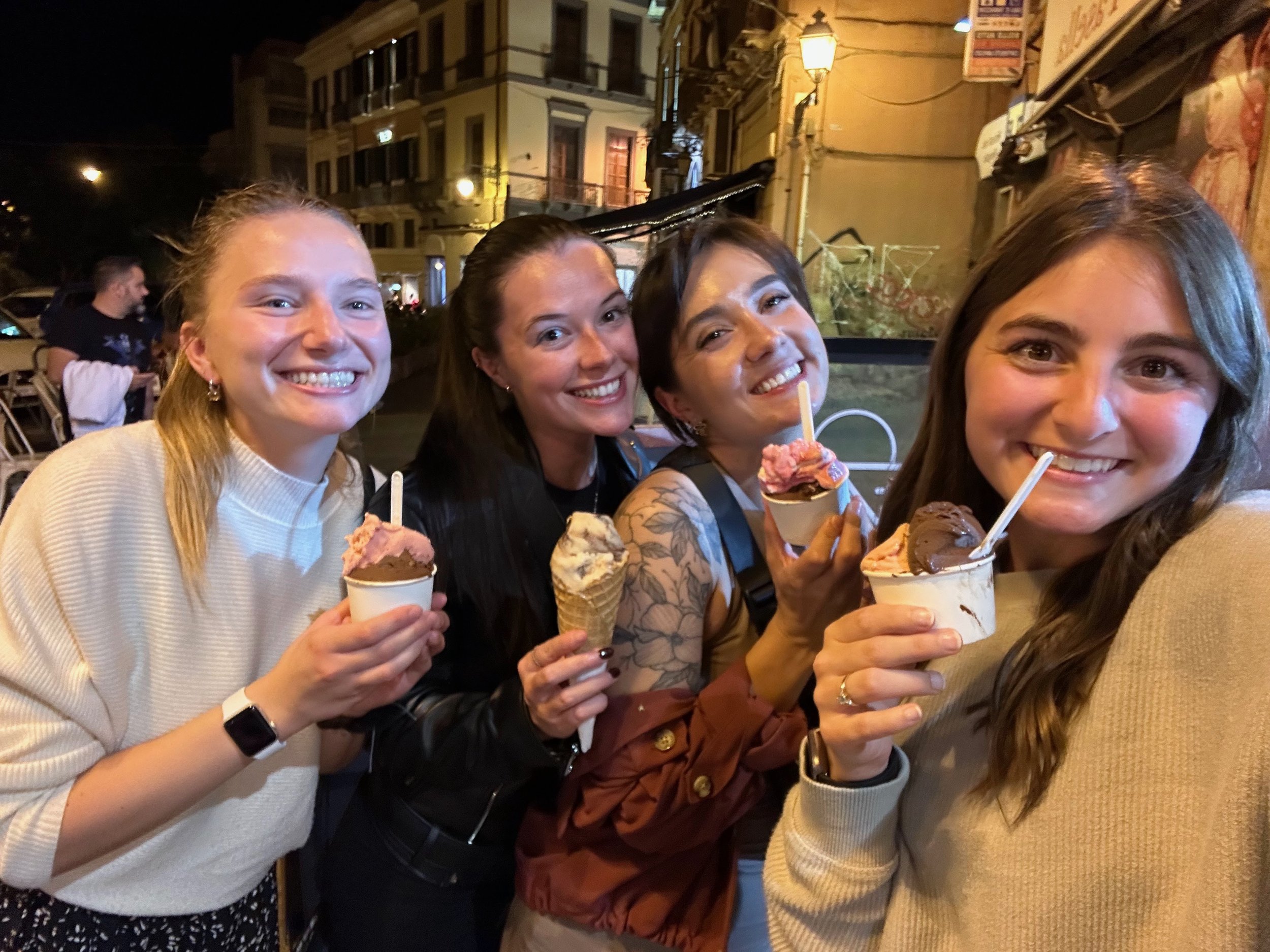 Four young women smiling and holding cups of ice cream at night outdoors, with city buildings and streetlights in the background.