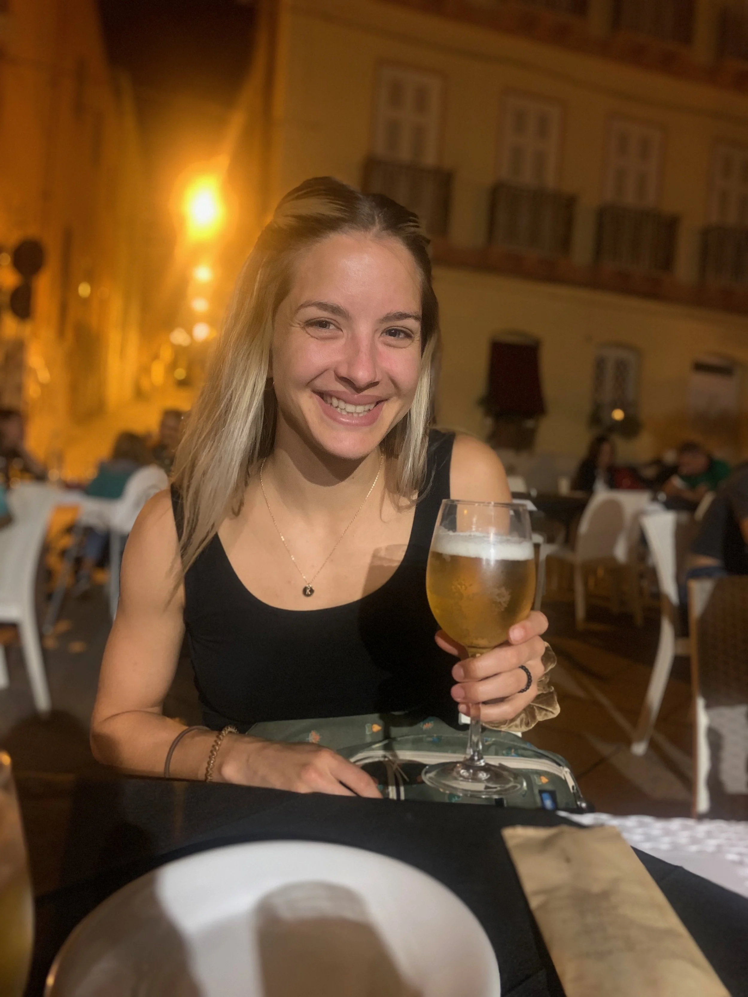 A young woman with blonde hair smiling and holding a glass of beer at an outdoor restaurant at night.