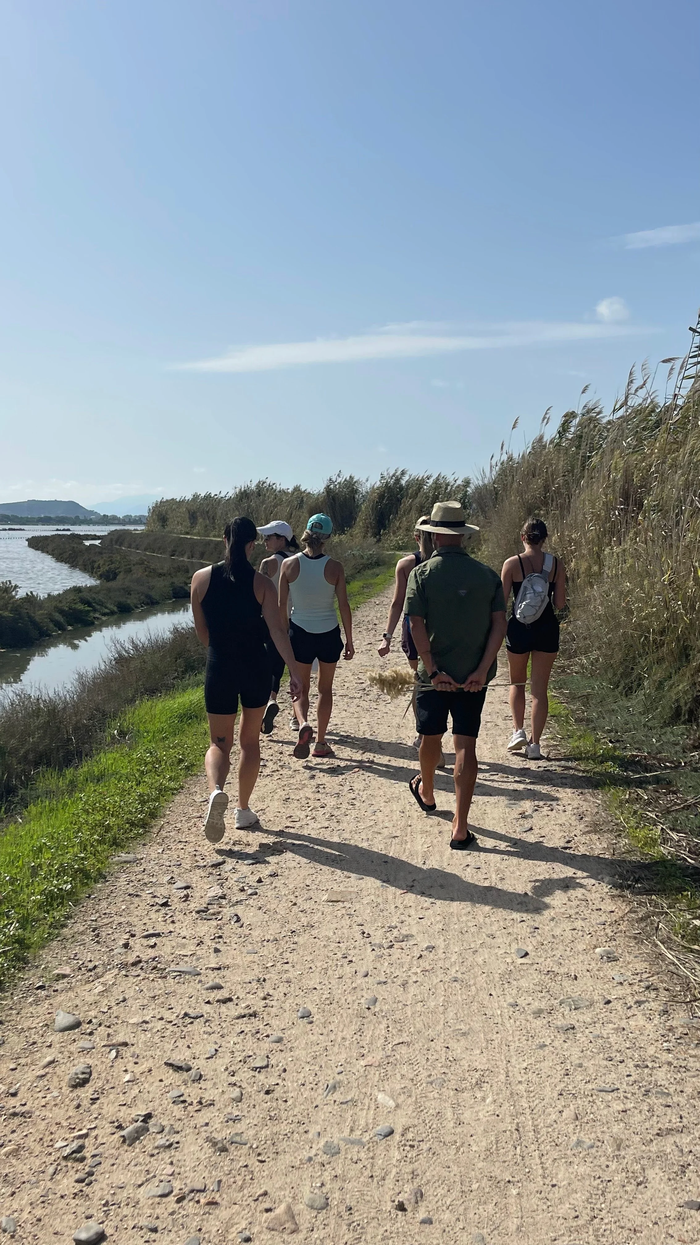 Group of seven people walking on a dirt trail beside a body of water, with tall grass and blue sky in the background.