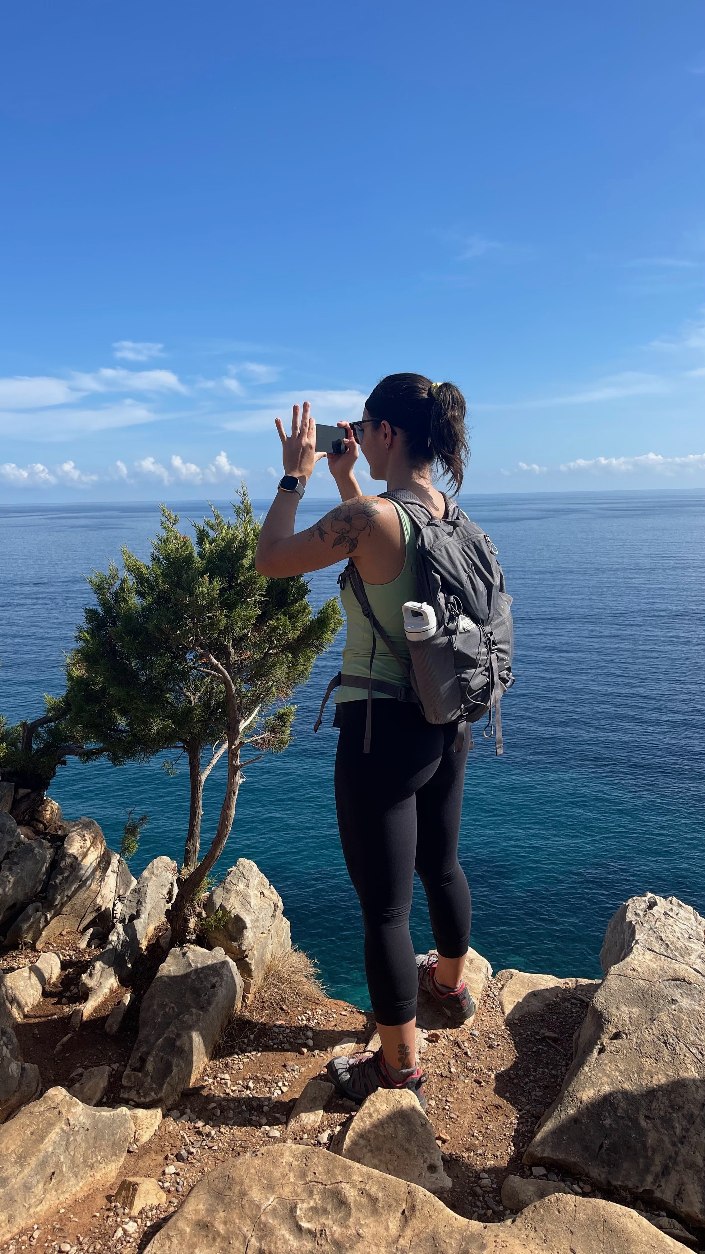 A woman with a backpack taking a photo with her phone on a rocky cliff overlooking the ocean under a clear blue sky.