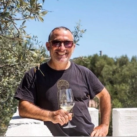 Sardalia tour guide holding a glass of white wine outdoors with trees and blue sky in the background.
