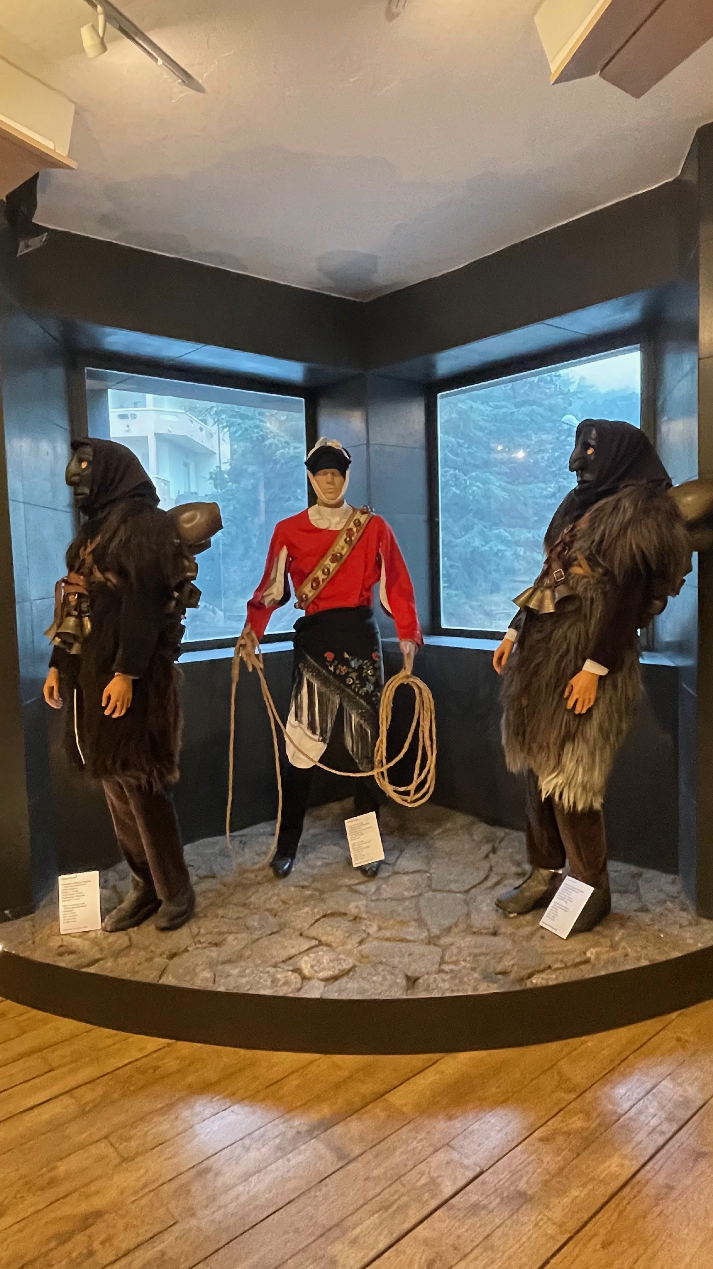 Two mannequins dressed in traditional Native American clothing with a third mannequin dressed in a red and black uniform holding a lasso, displayed in a museum exhibit with windows showing trees outside.