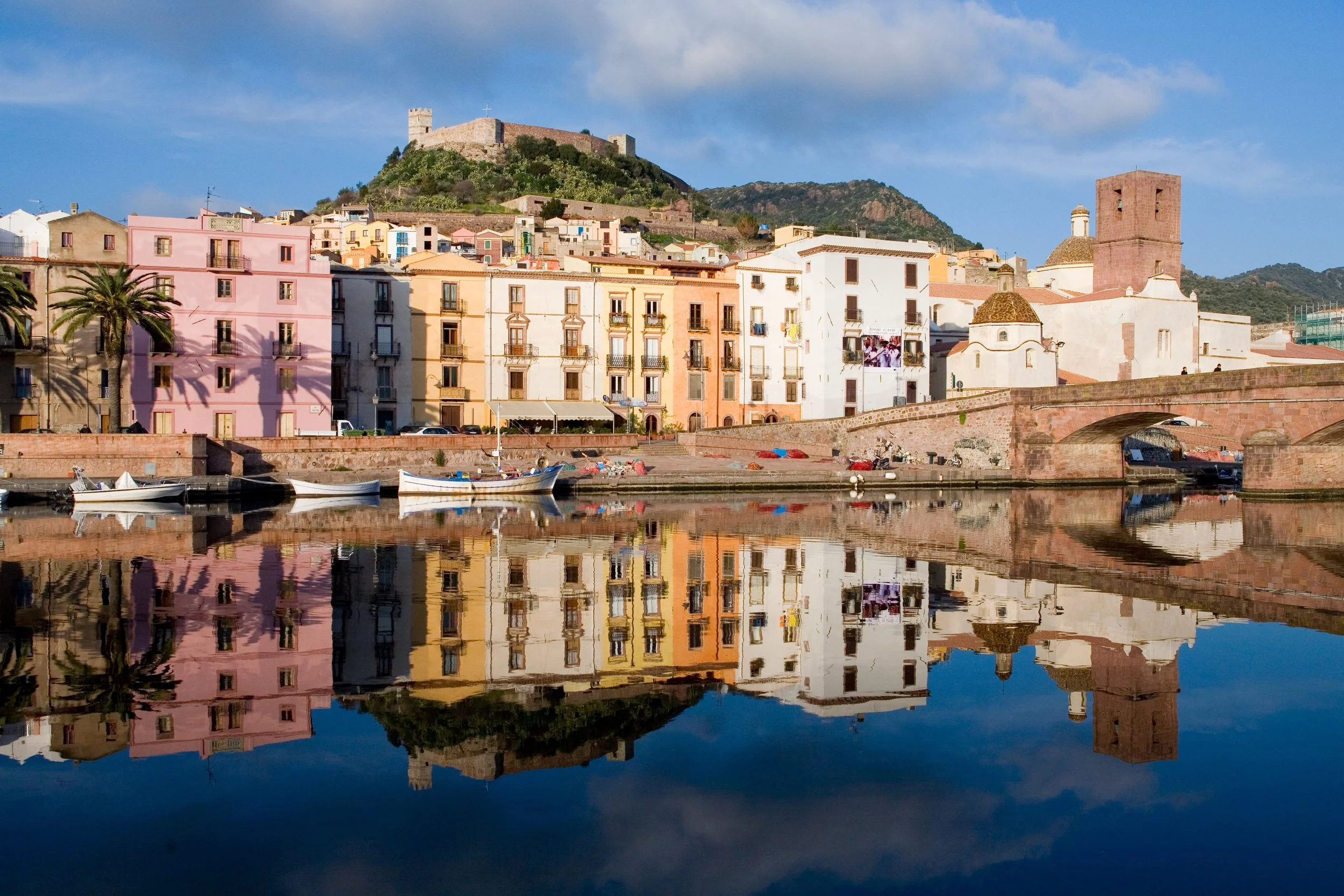 Bosa, Sardinia - Colorful buildings along a waterfront with boats, reflected in calm water, hillside castle in background, blue sky with clouds.