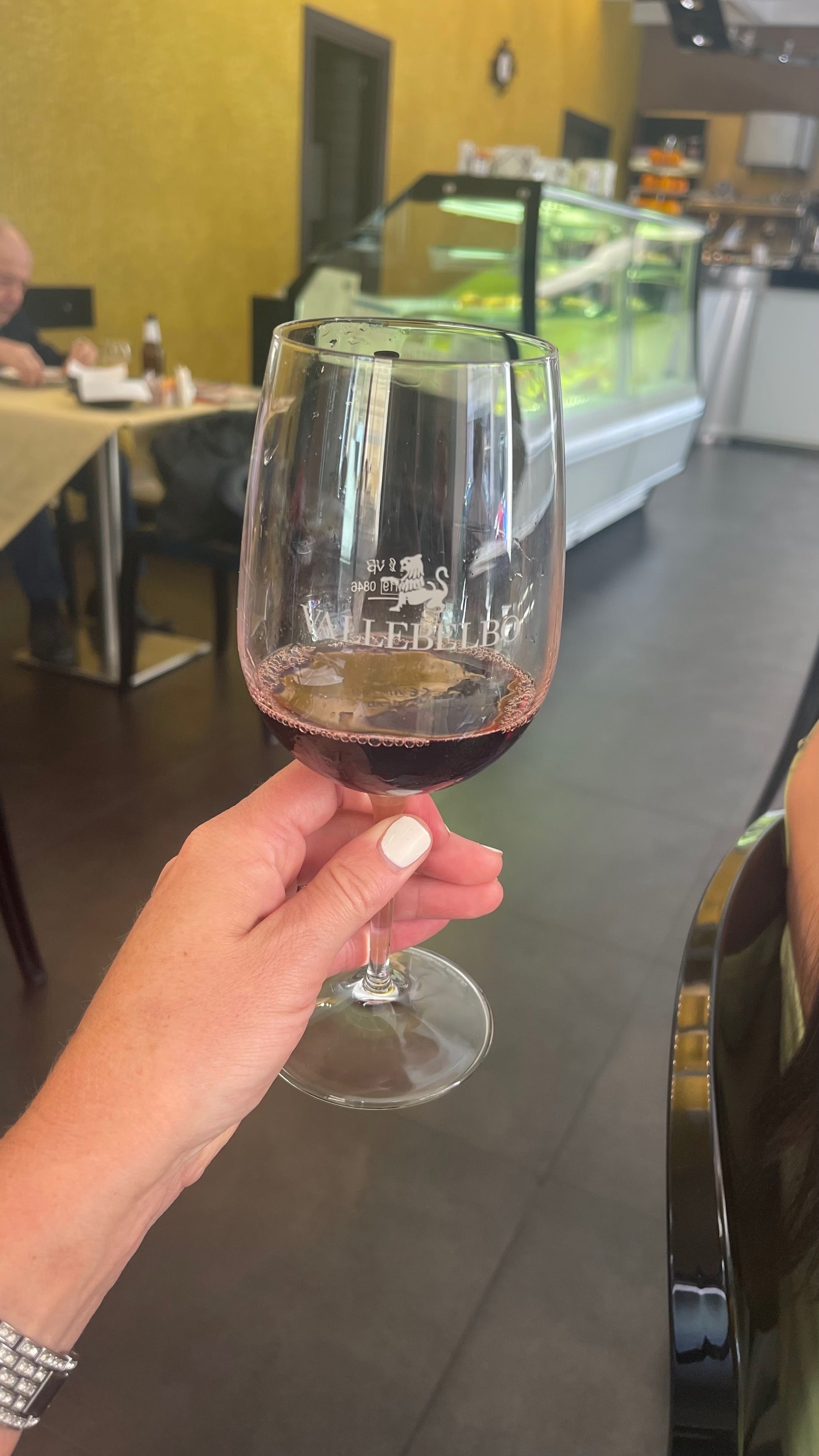 A person holding a glass of red wine inside a restaurant or cafe, with a yellow wall, a glass display case, and a customer in the background.
