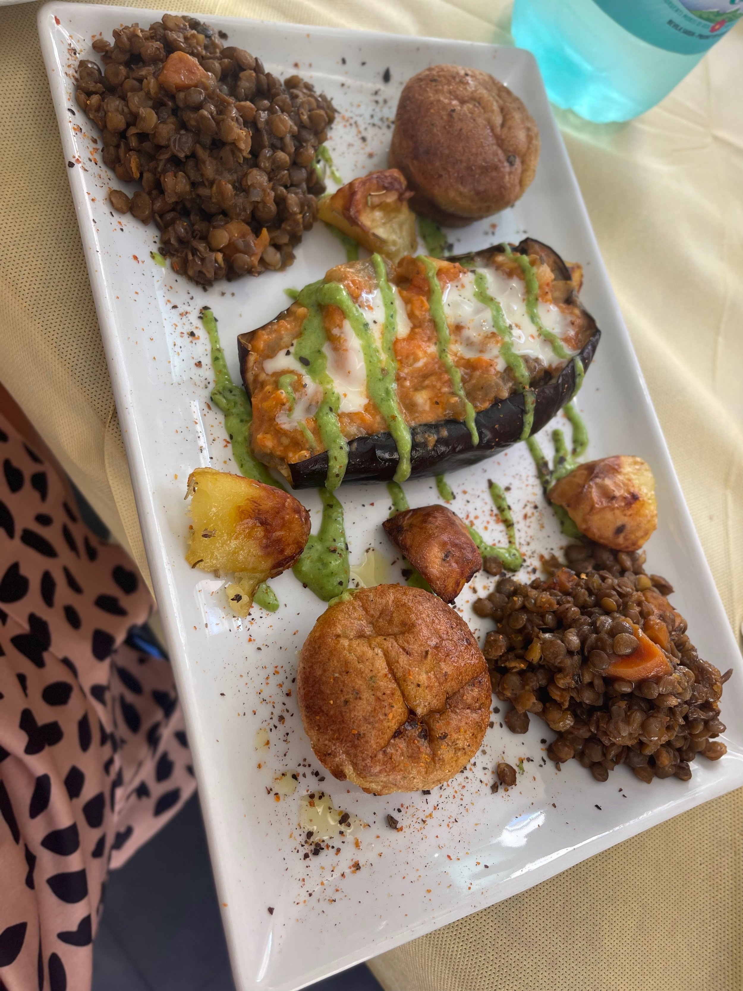 A white rectangular plate with baked eggplant topped with white sauce and green sauce, surrounded by lentils, roasted potatoes, and fried plantains. There are two bread rolls on the plate and a blue beverage in a plastic bottle in the background.
