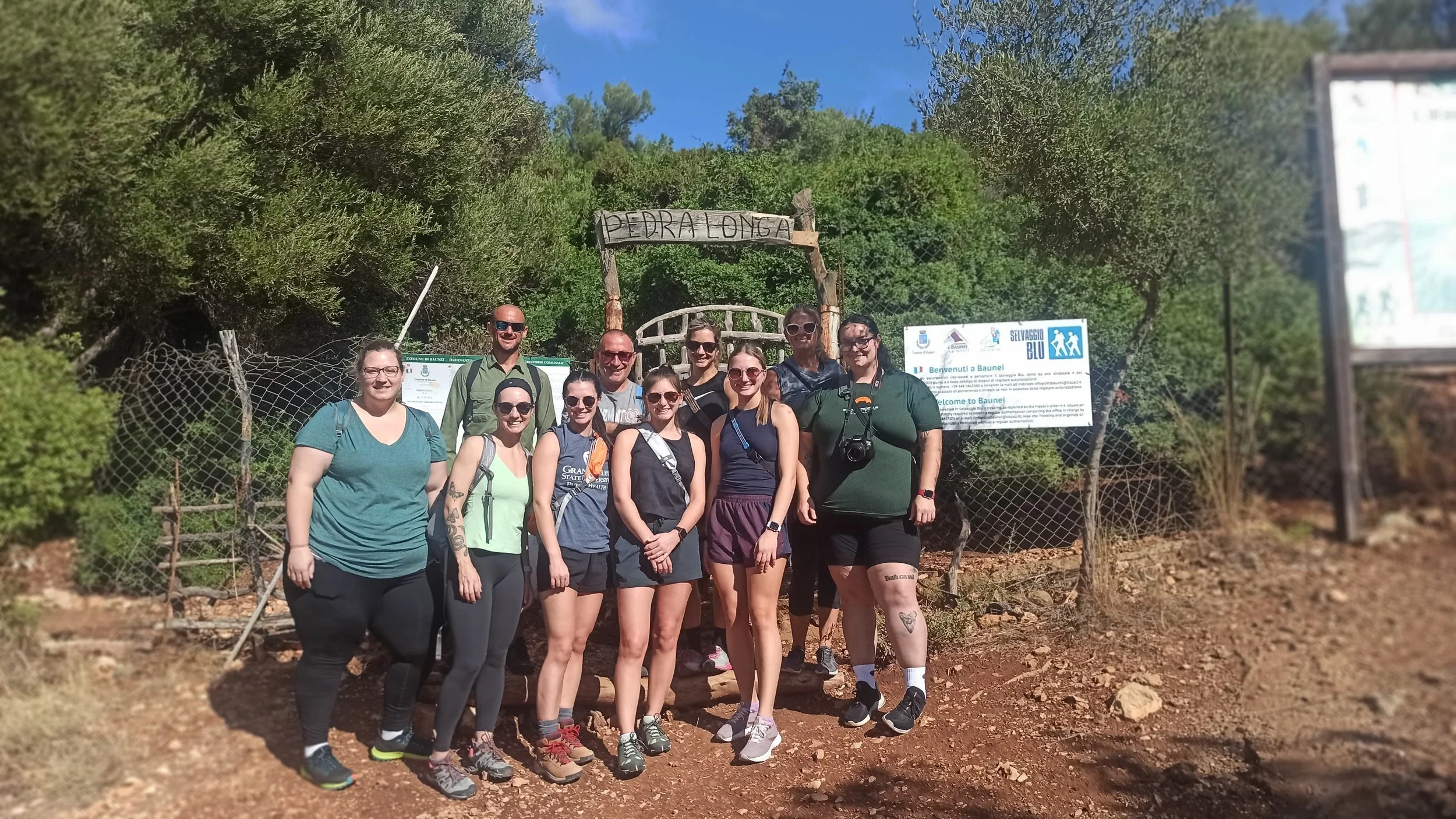Group of ten people posing outdoors in front of a wooden sign that says 'PEDRALONGA.' They are on a hiking trail with green bushes and trees behind them, and informational signs to the right.