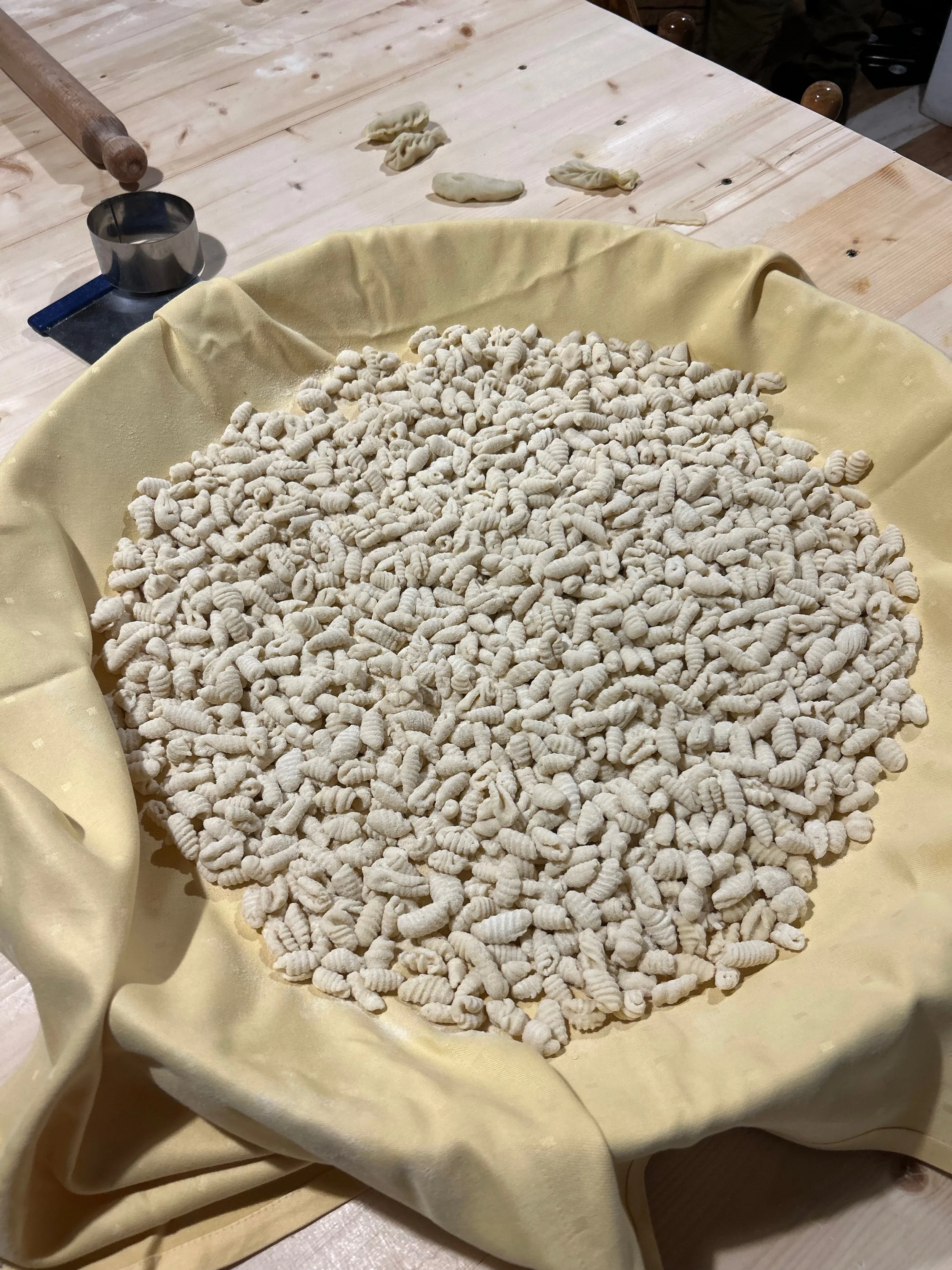 A large pile of small, white, freshly made pasta pieces on a yellow cloth on a wooden table.
