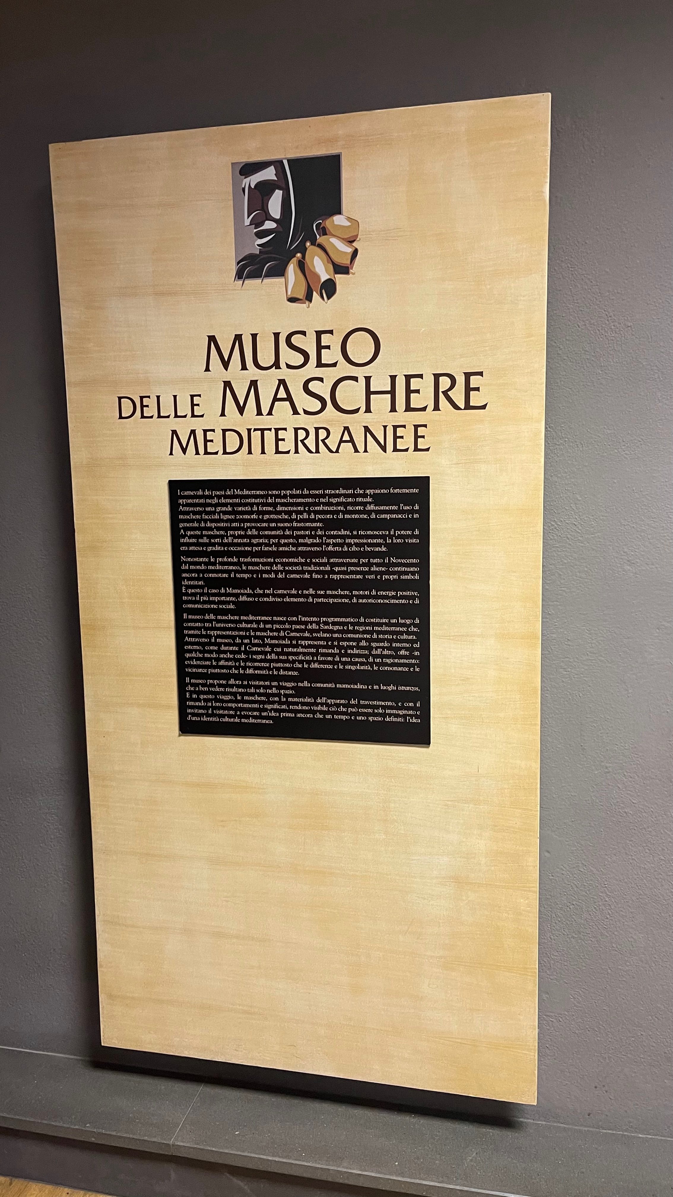 Sign for the Museum of Masks of the Mediterranean, featuring a logo of a mask and bells at the top. The sign contains descriptive text about the history and cultural significance of masks in the Mediterranean region.