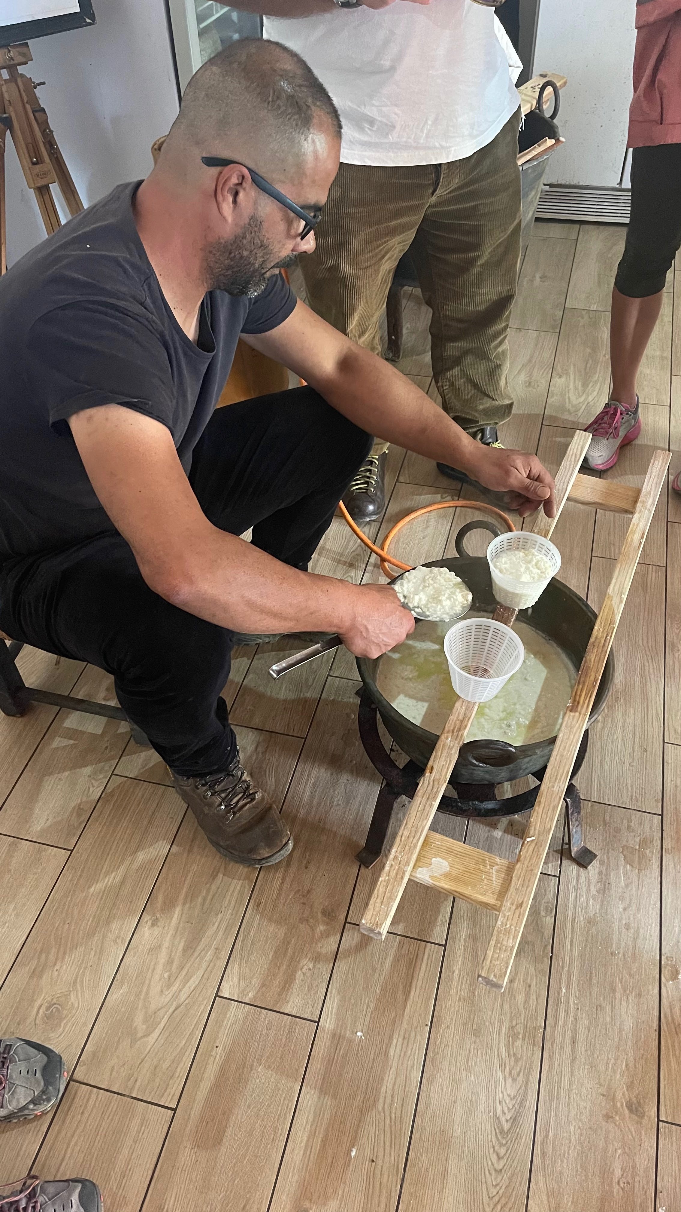 A man sitting on a chair using a ladle to scoop cheese from a large hot pot, with a funnel and a small basket nearby, on a wooden floor inside a restaurant.