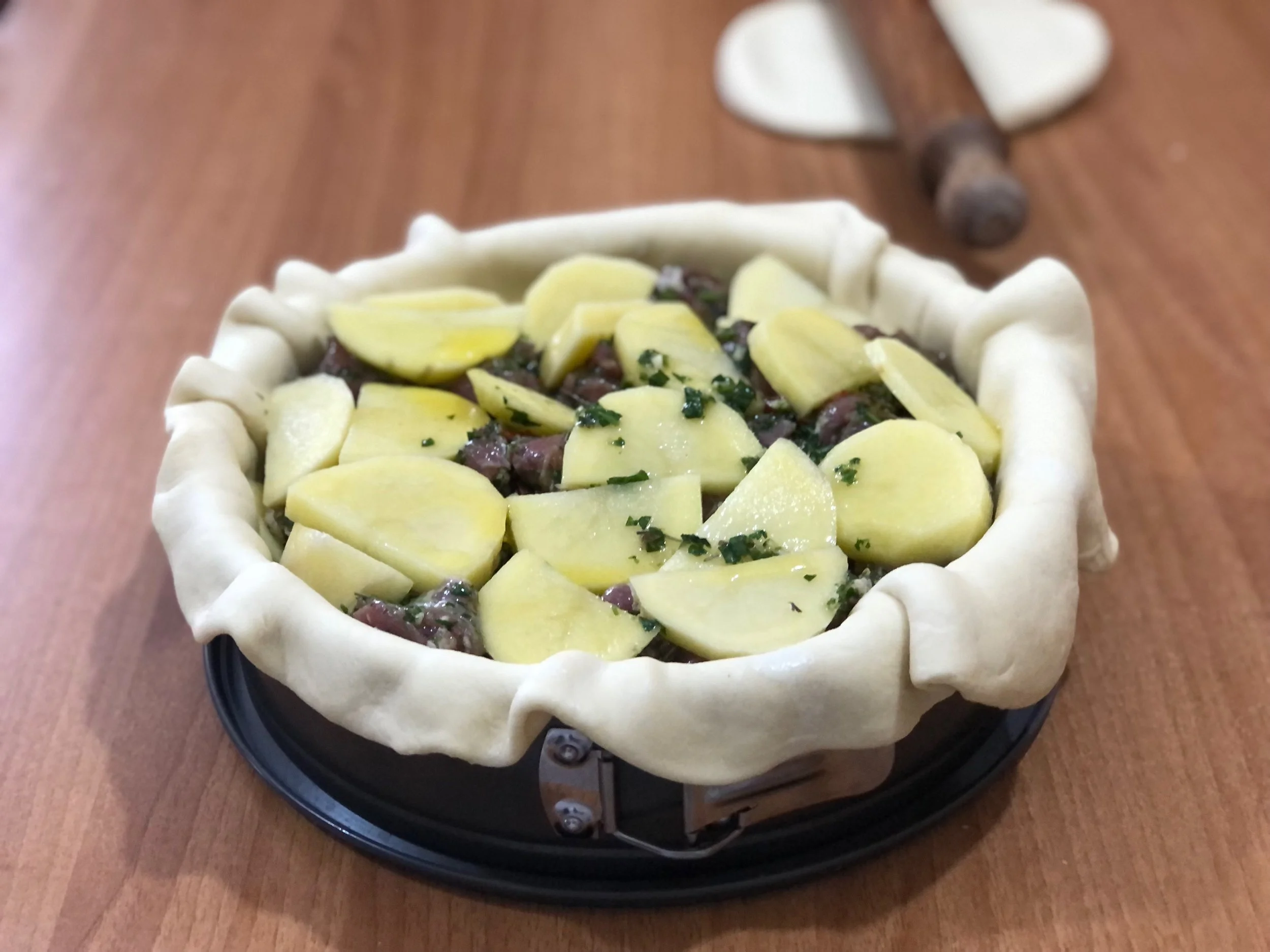 Unbaked pie crust filled with sliced potatoes, chopped herbs, and other ingredients on a round baking pan.
