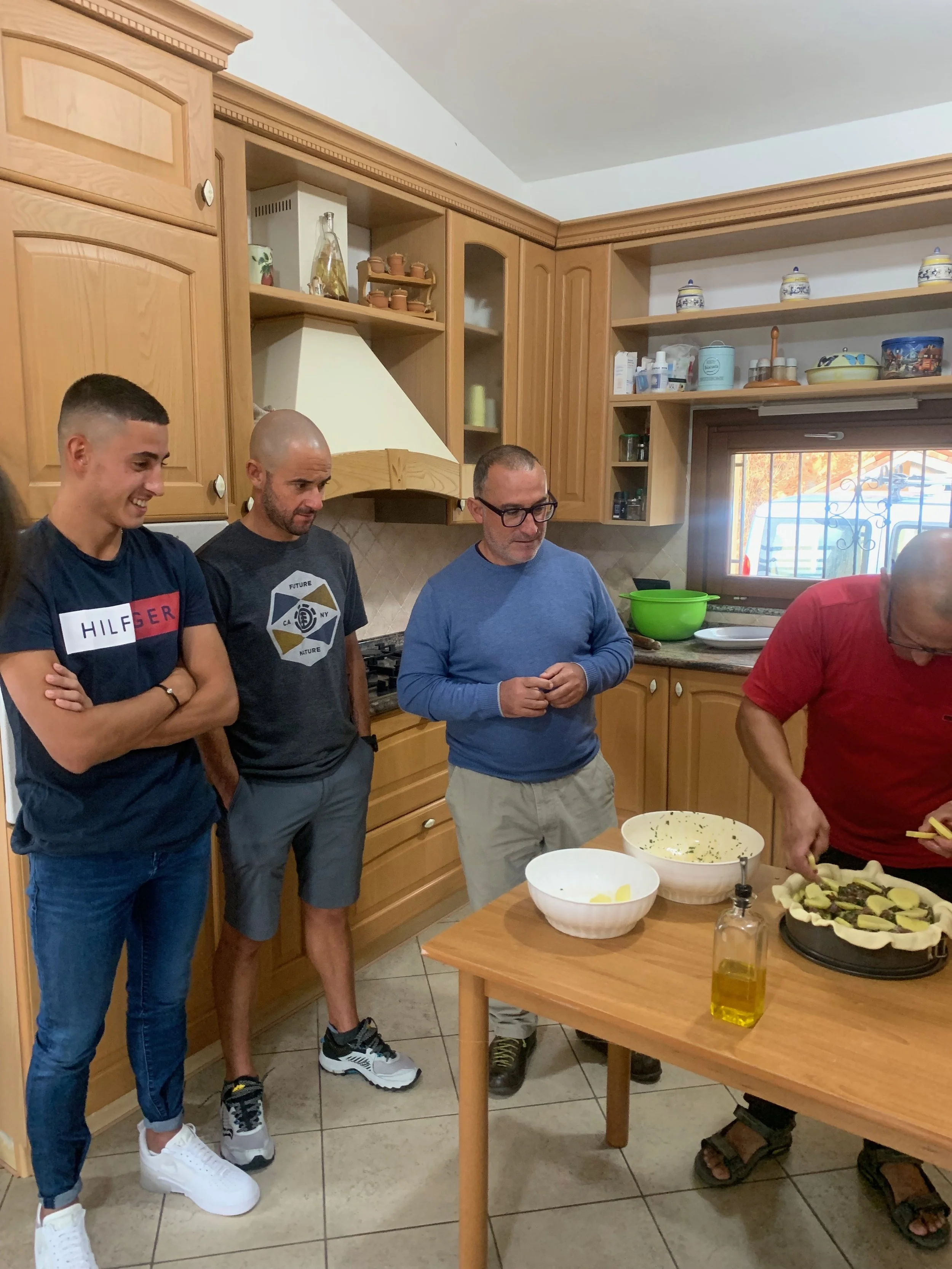Four people in a kitchen, one preparing food on a table, while others watch. The kitchen has wooden cabinets and a window with bars.