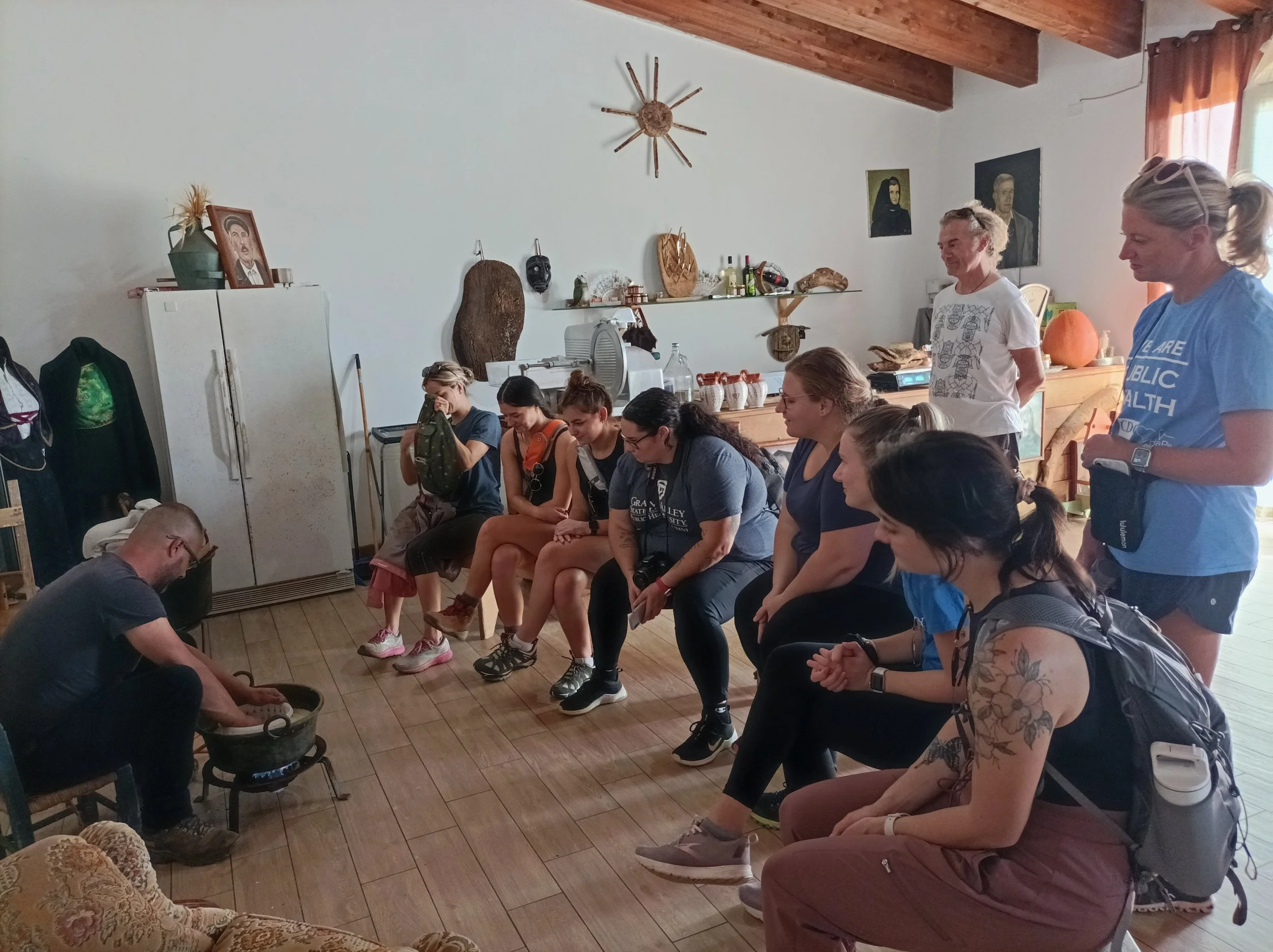 Group of people seated in a room watching a man demonstrate grinding something in a bowl over an open flame.