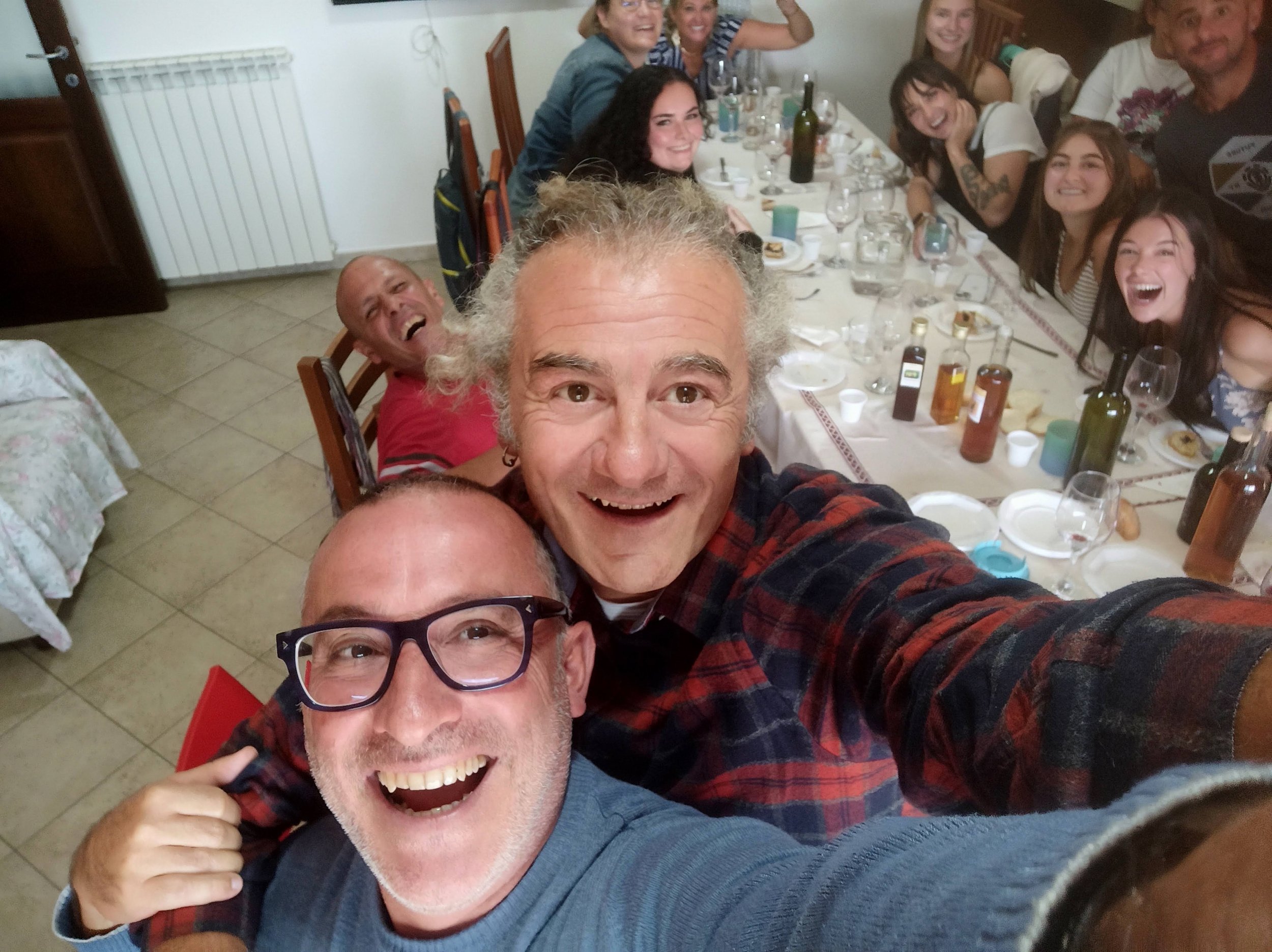 Group of people gathered around a dinner table, smiling and taking a selfie with two men closest to the camera. The table has plates, glasses, bottles, and food items, and the room appears to be a cozy dining area.