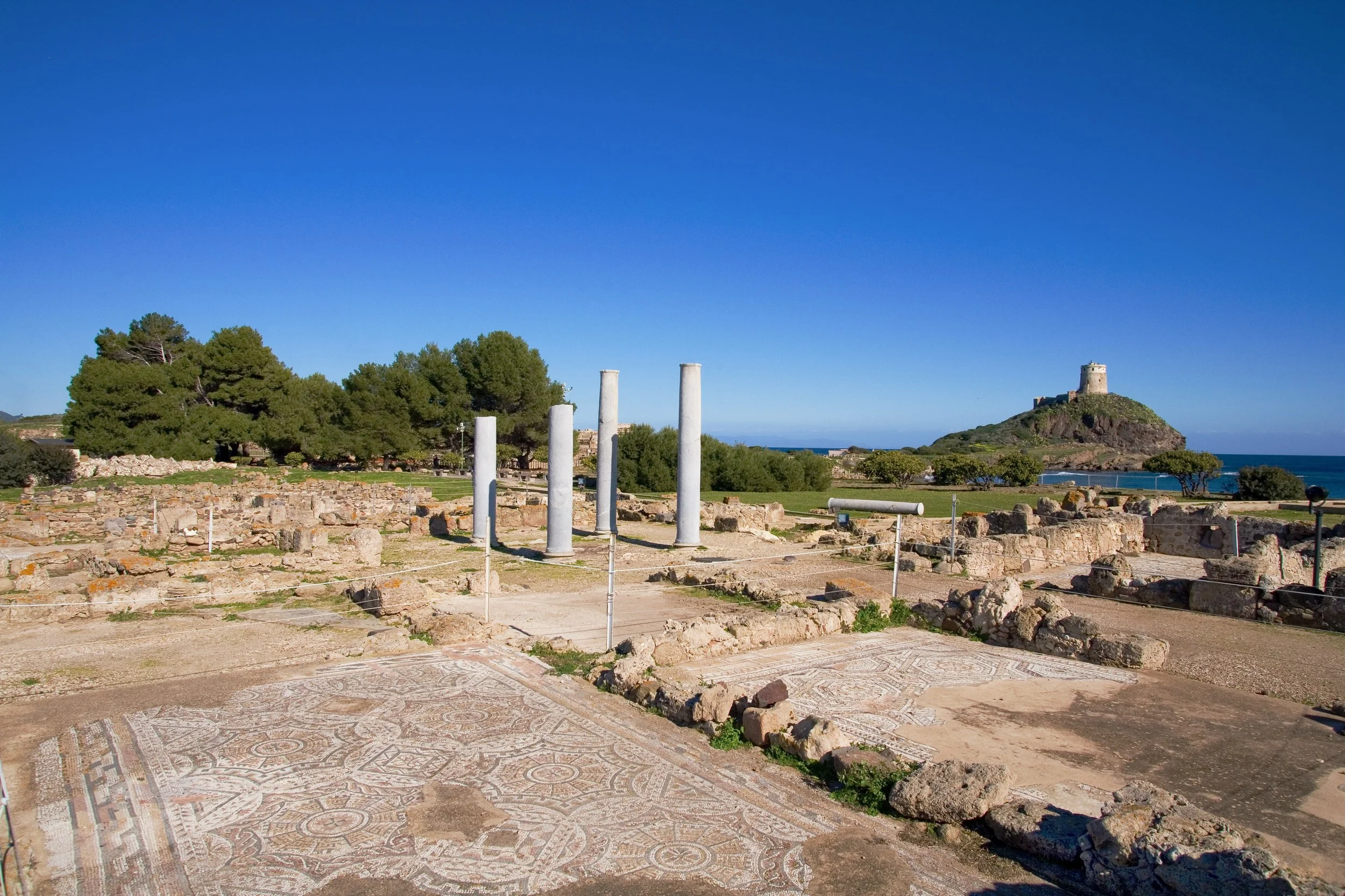 Nora, Sardinia - Ancient Roman ruins with standing columns, mosaic floors, and a ancient Pisan-built look-out tower on a hill in the background, under a clear blue sky.