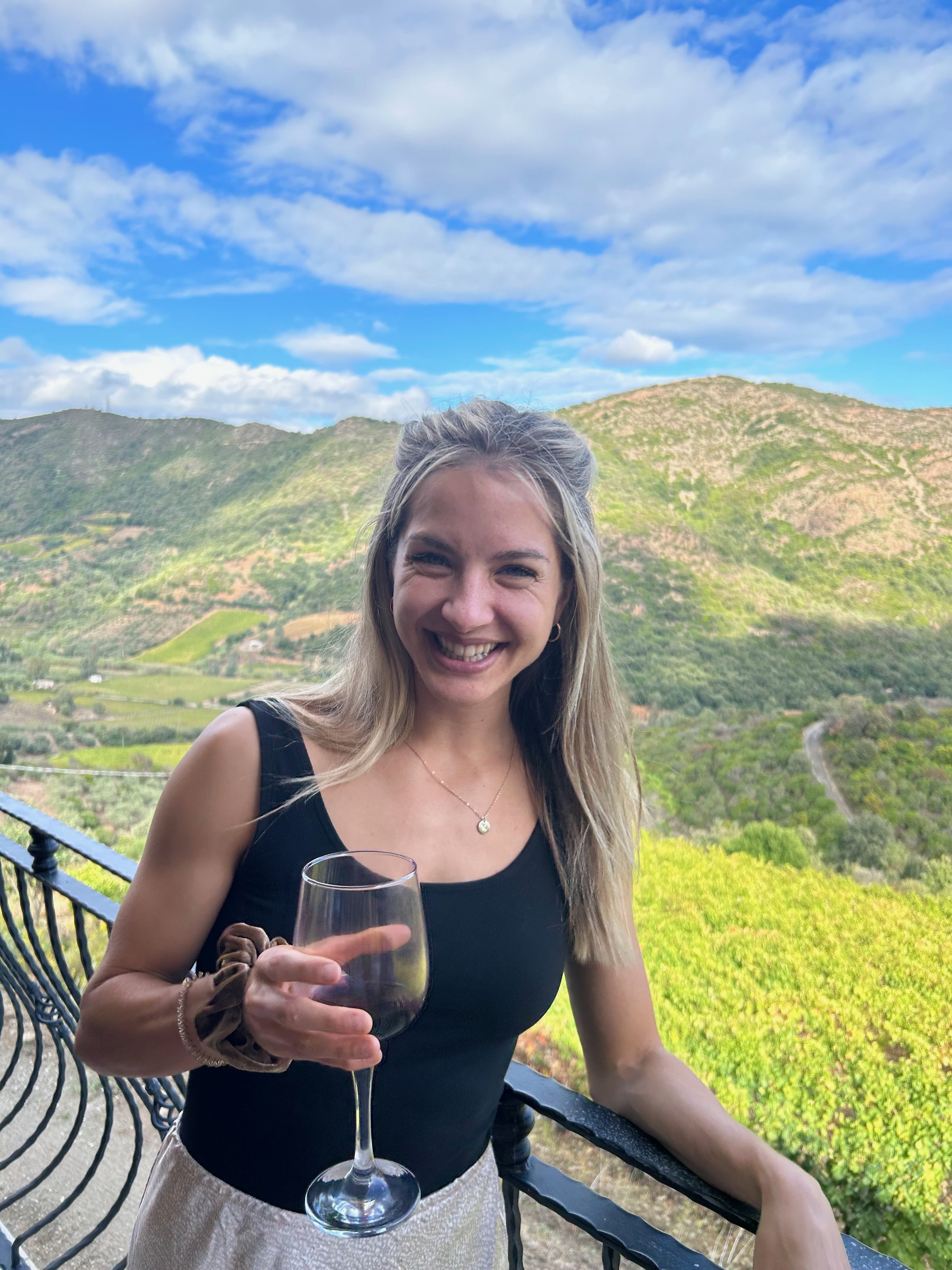Smiling woman holding a glass of red wine on a balcony with a lush green hillside and blue sky in the background.