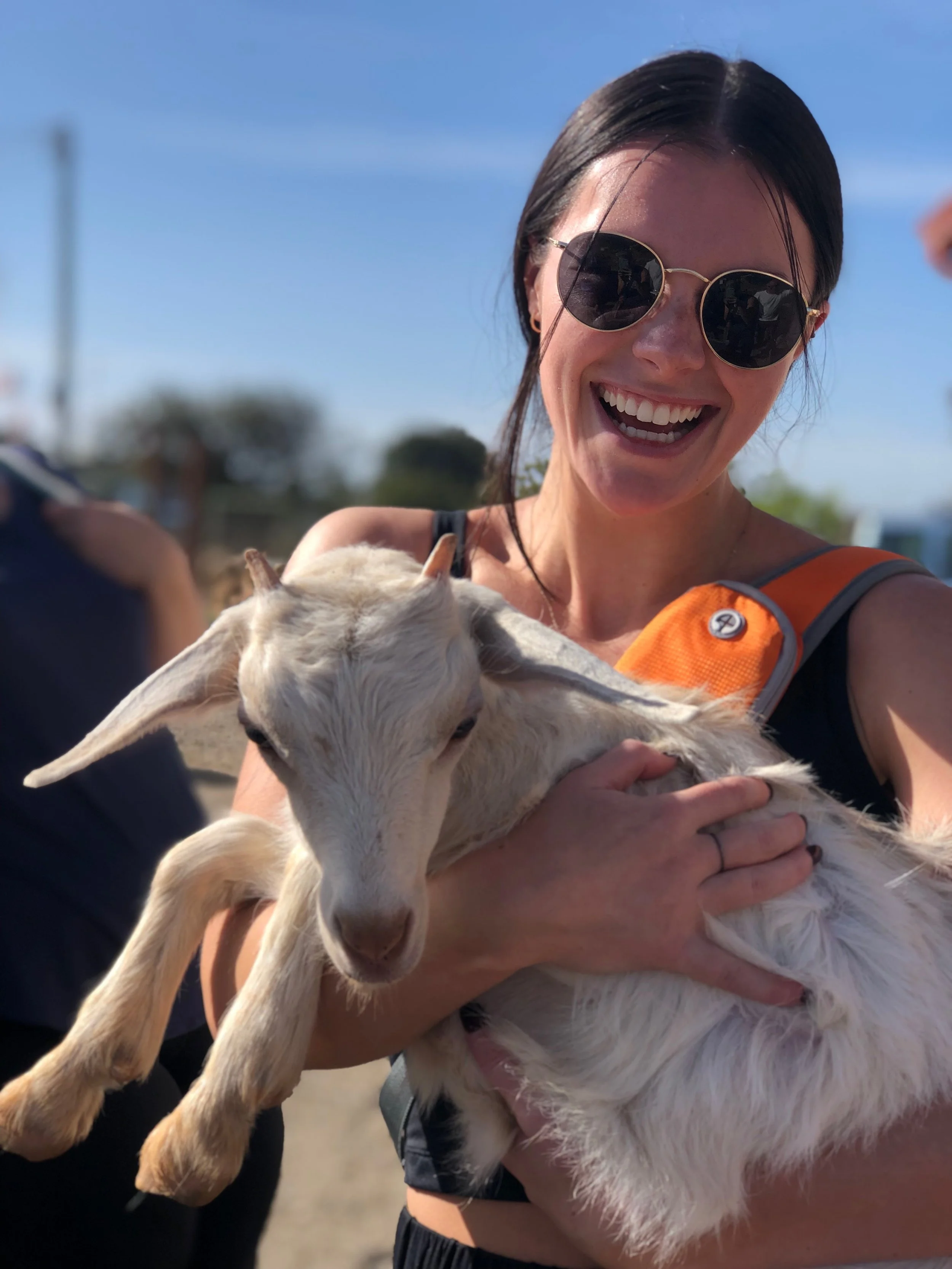A smiling woman wearing sunglasses holding a small white goat in her arms outdoors on a sunny day.