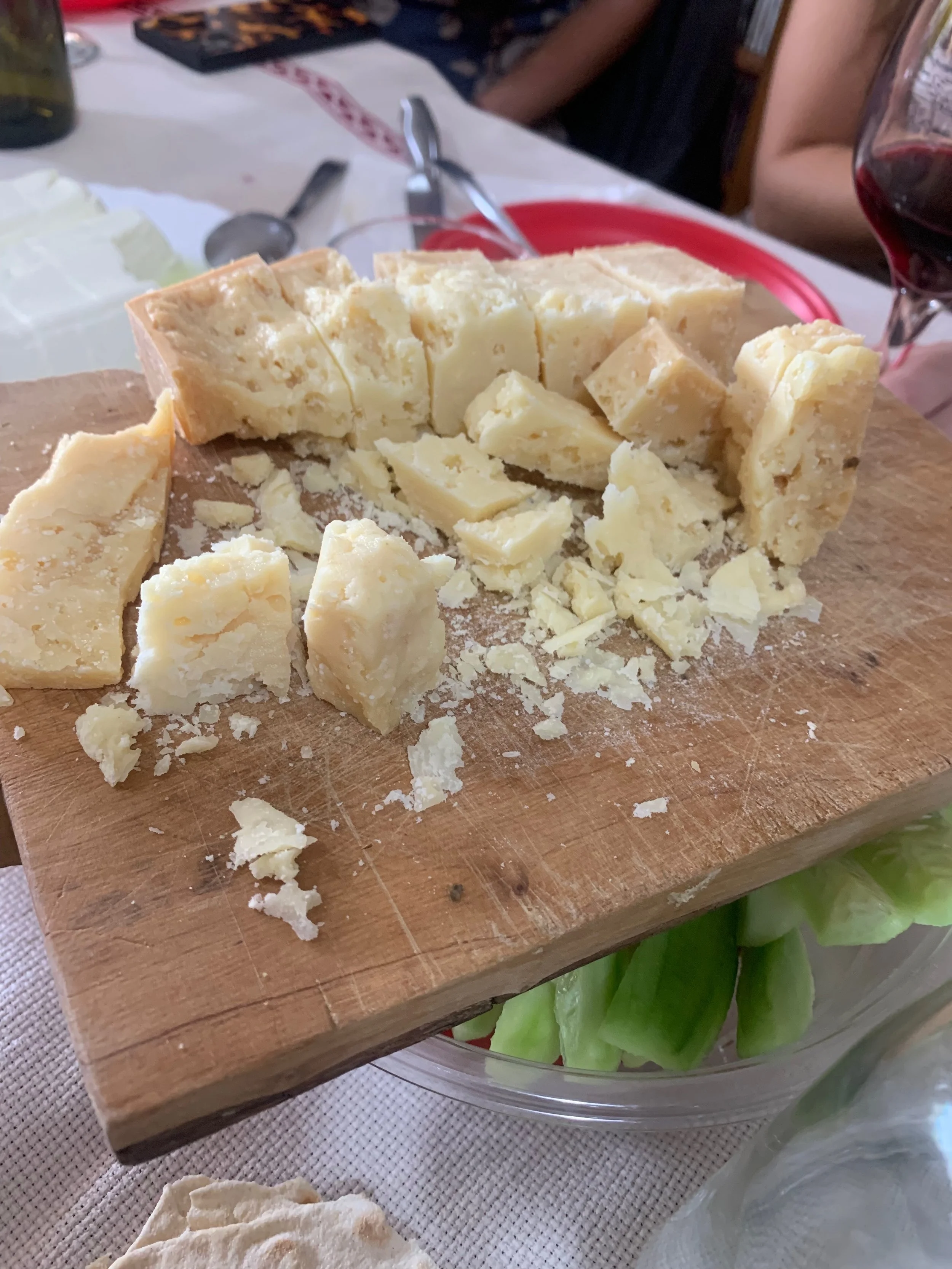 A wooden cheese board with chunks of hard cheese on it, placed over a glass bowl of sliced vegetables, possibly cucumbers.