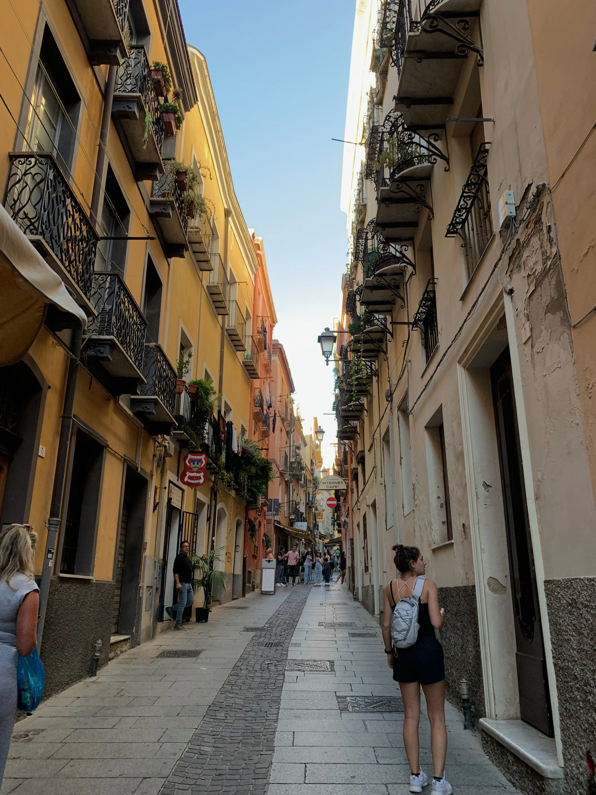 A narrow cobblestone street in a European city, lined with colorful buildings with balconies, some with plants. People walk along the street during daytime under a clear sky.