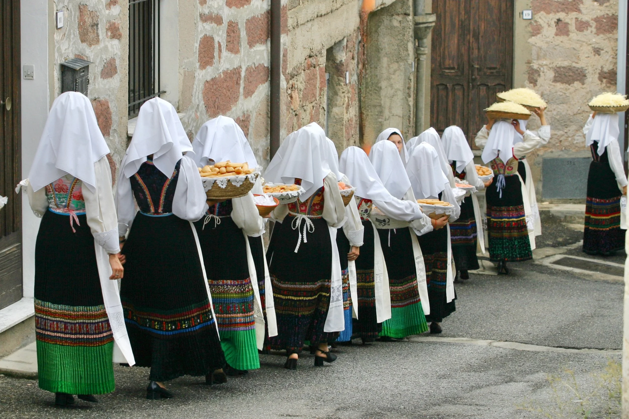 A group of Sardinian women in traditional costumes walking along a stone-paved street, carrying trays and baskets of bread, with some women wearing white veils over their heads.