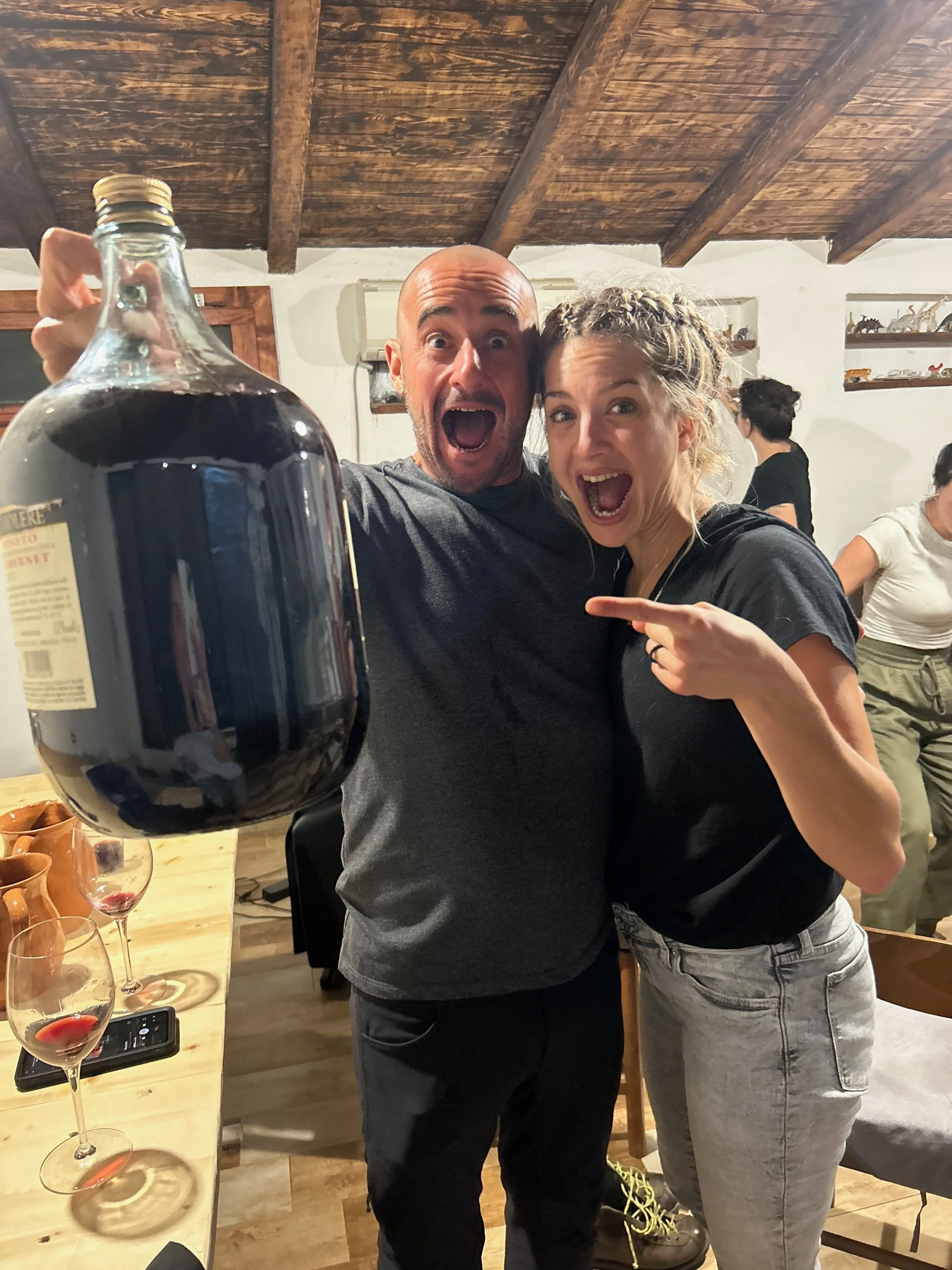 Two people, a man and a woman, are excitedly posing for a photo at a gathering, with the man holding a large bottle of dark liquid and both of them with expressive faces.