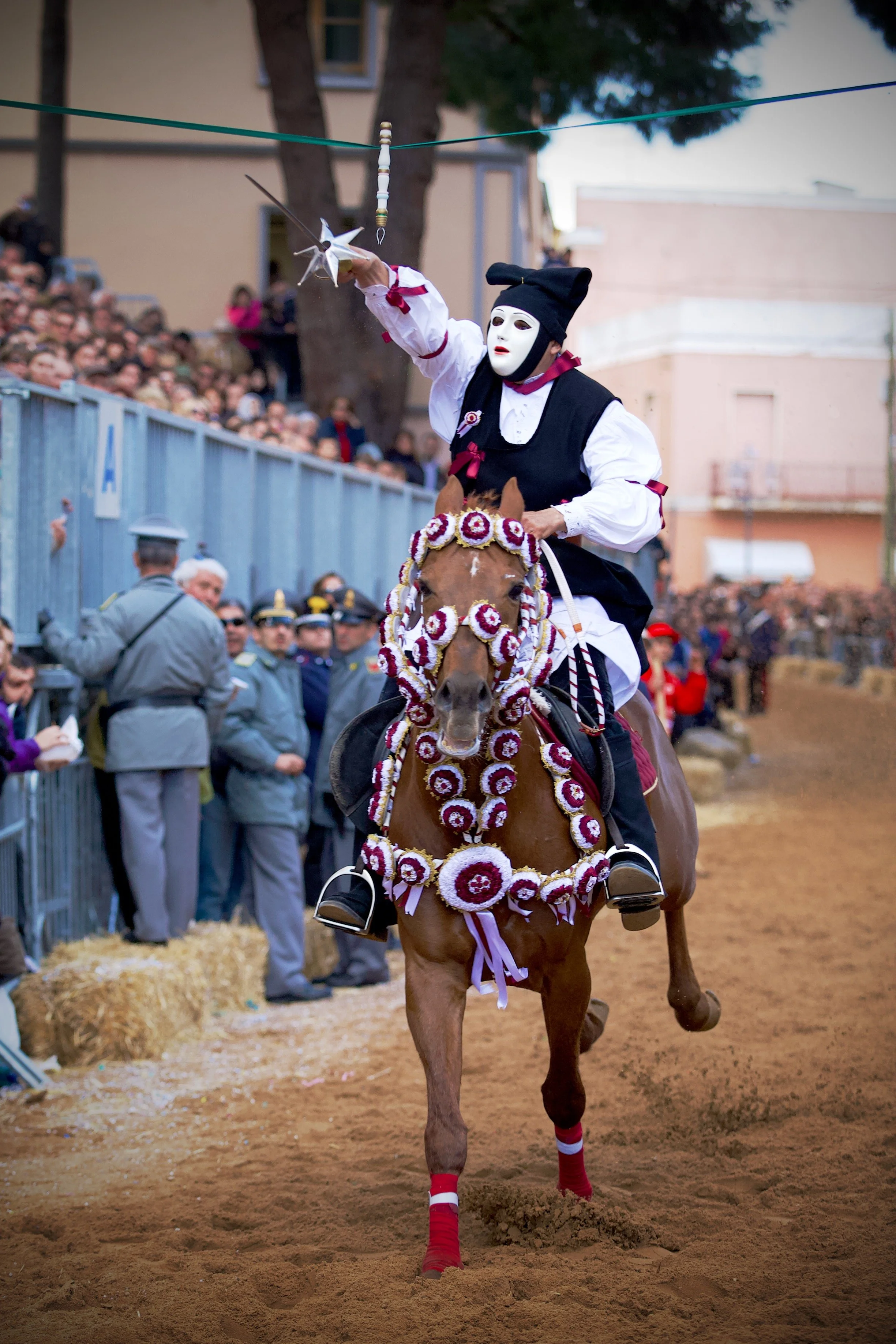 A Sardinian horseman dressed in traditional Sartiglia costume, grasping a sword with a white mask and black outfit riding a decorated horse during the Sartiglia festival. The horse is adorned with floral decorations.