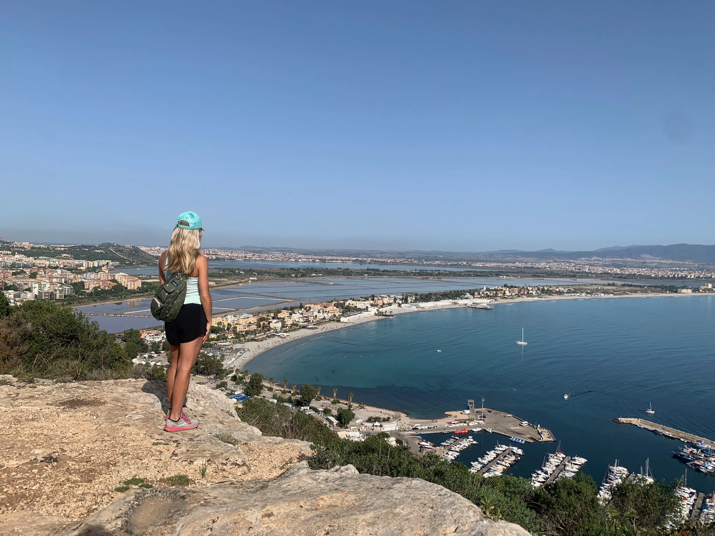 A woman standing on a rocky hill overlooking a coastal city with a marina and water bodies, under a clear blue sky.