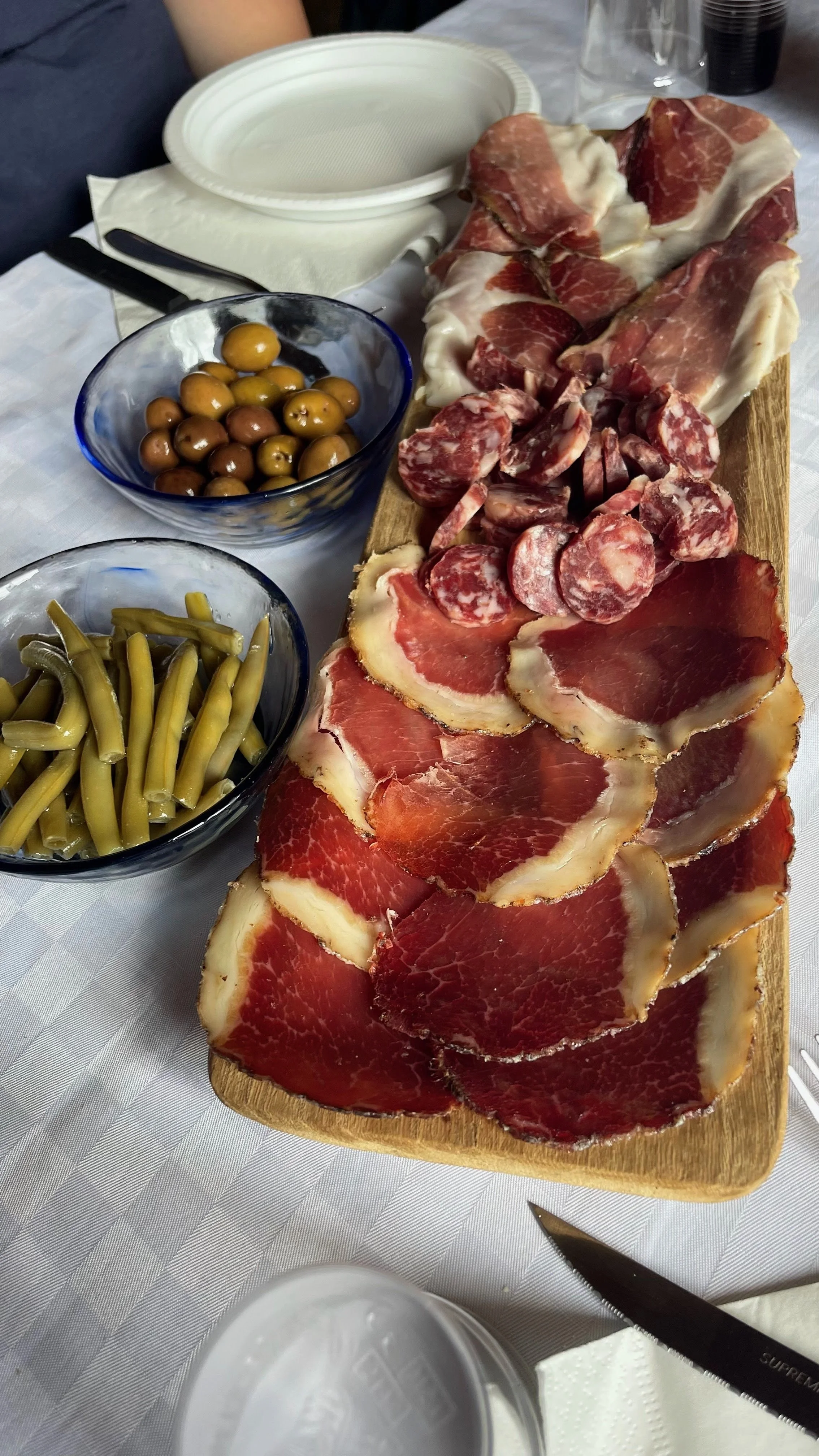 A wooden board with slices of cured meats and sausages beside bowls of green beans and olives on a white tablecloth.