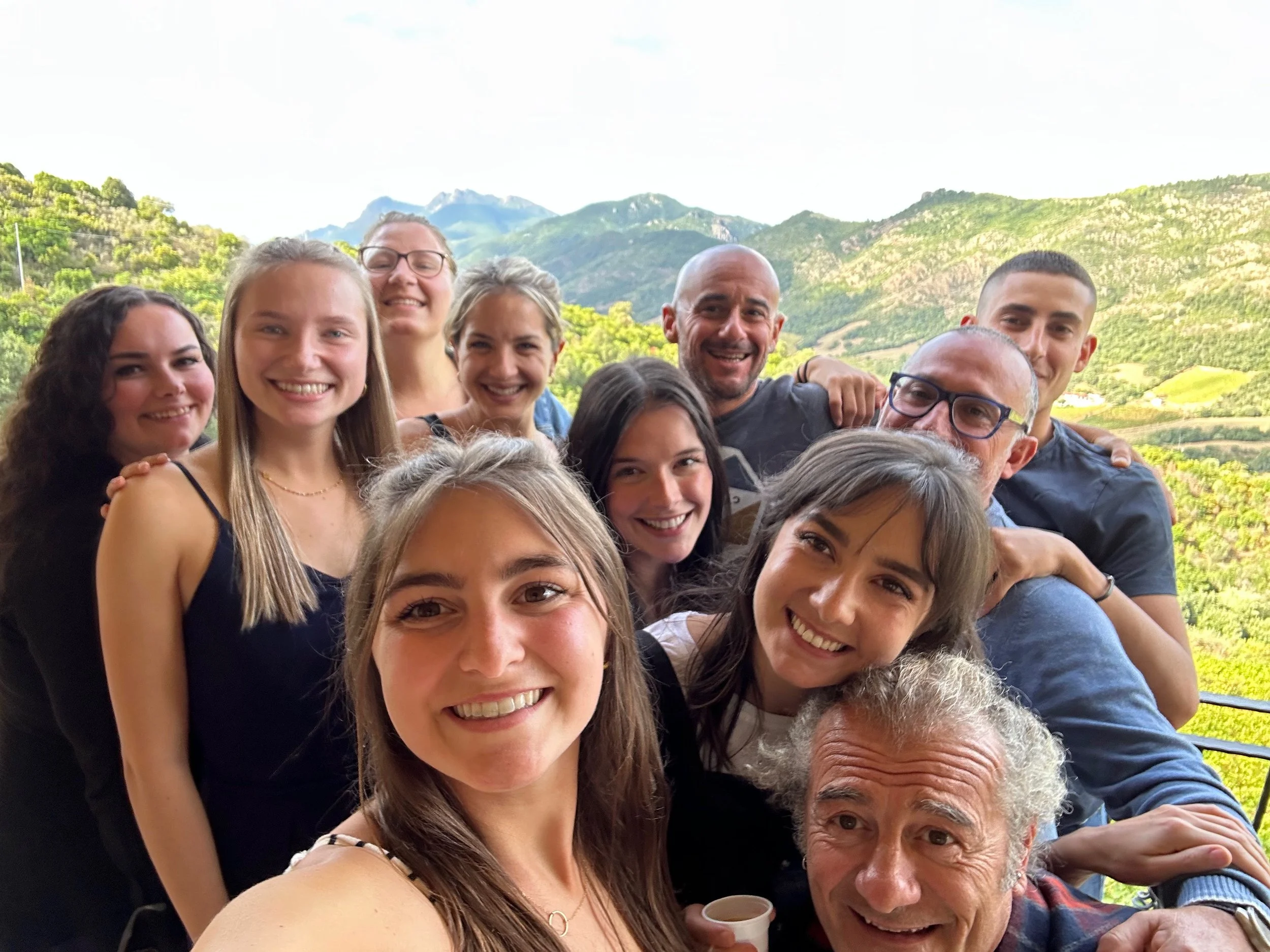 Group of ten people smiling and posing for a selfie outdoors with green mountains in the background.