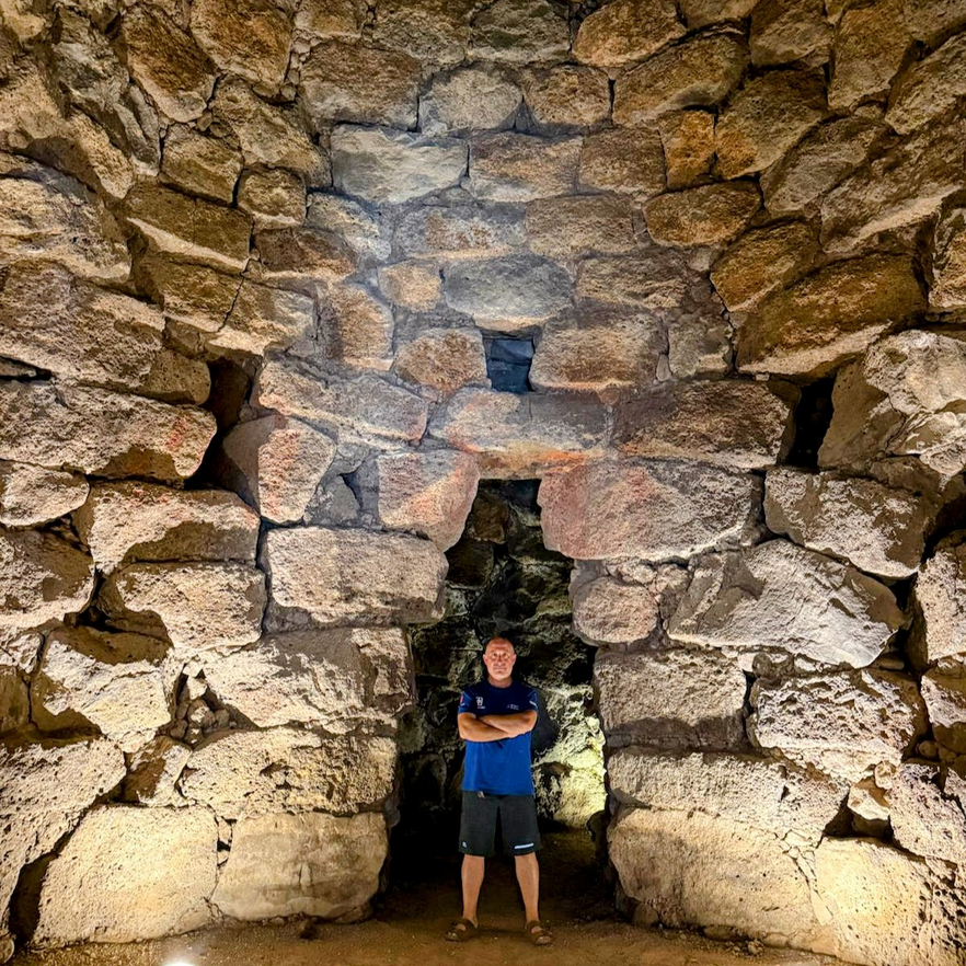 A man standing with arms crossed inside an ancient stone structure called Nuraghe in Sardinia, with large rough-hewn stones forming arches and walls.