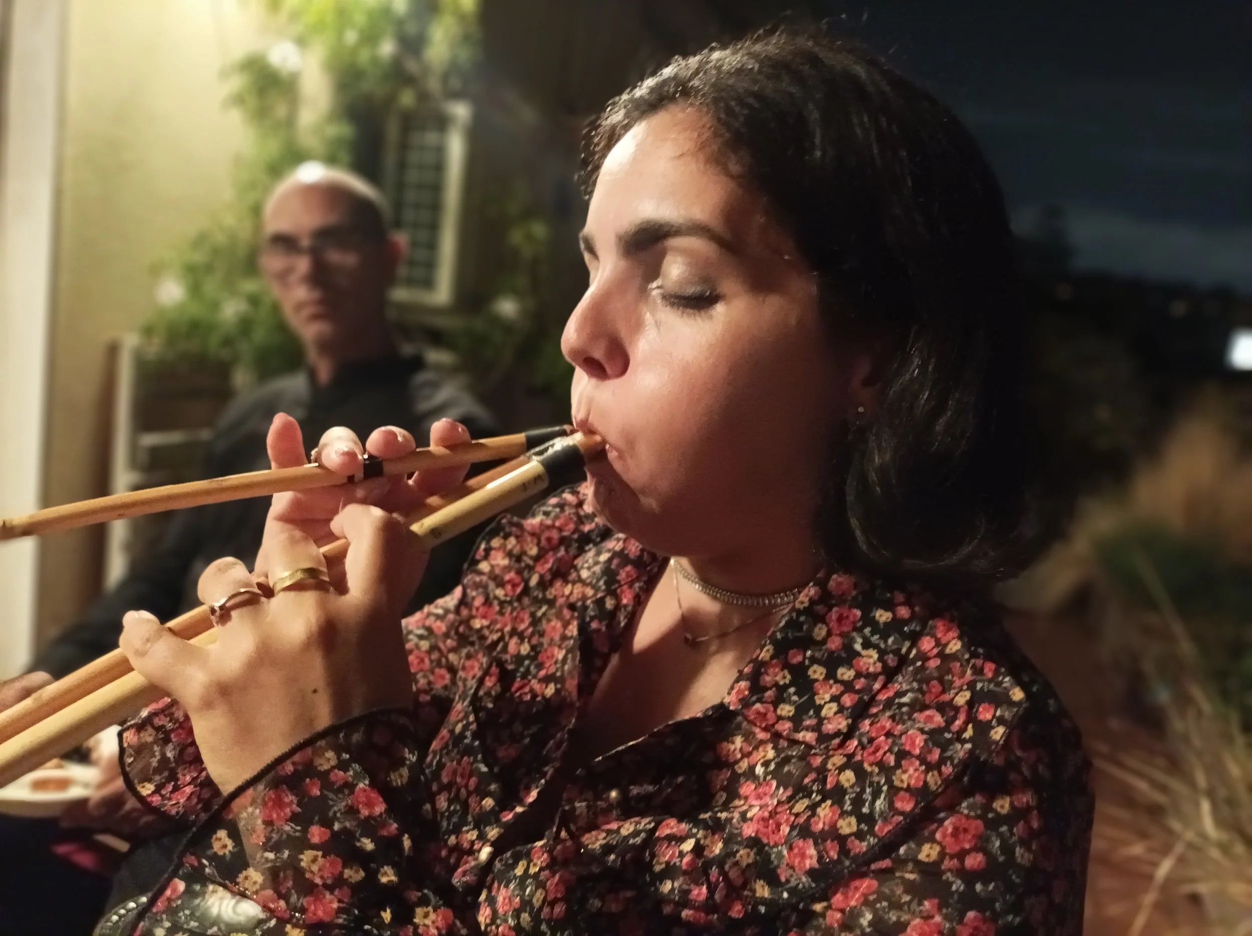 A woman with dark hair playing a traditional wooden flute, with a man in glasses blurred in the background under warm lighting at night.