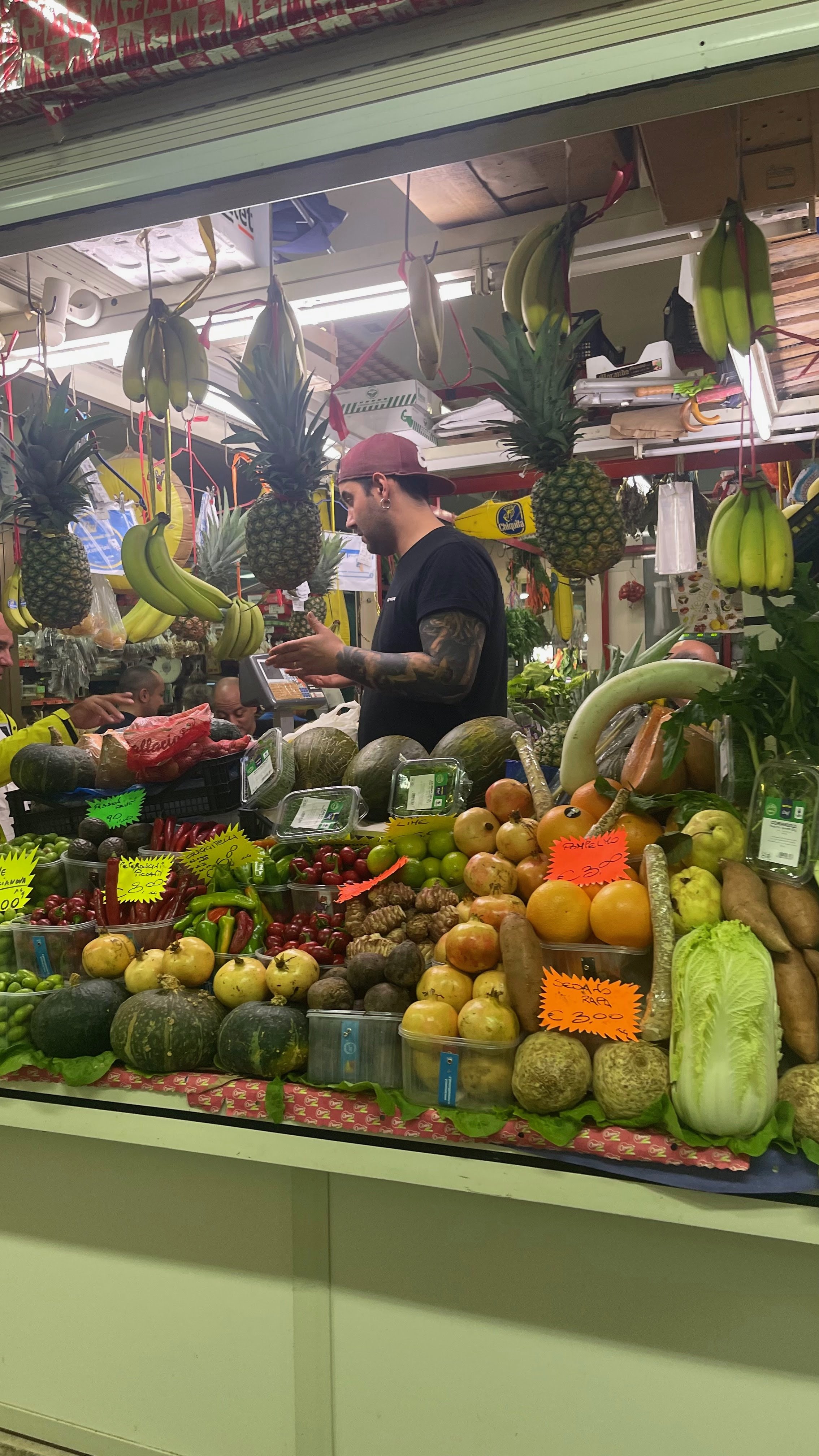 A fruit market stall displaying pineapples hanging upside down, bananas, pumpkins, apples, oranges, and other fresh produce. A man with tattoos is at the checkout counter talking to a cashier.