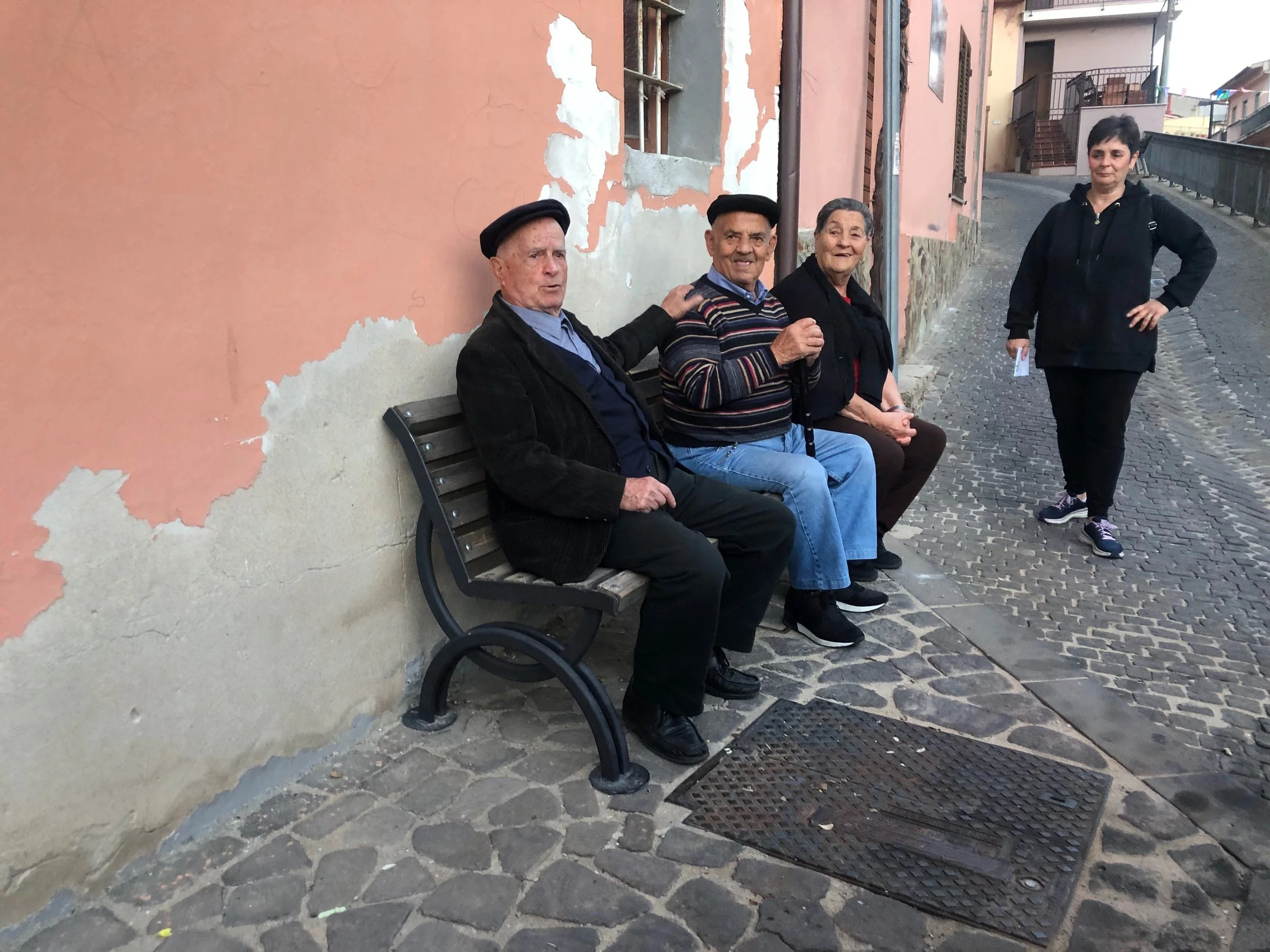 Four people sitting and standing on a cobblestone street near a weathered building with peeling paint. Three older adults are seated on a bench, and a younger woman is standing nearby.