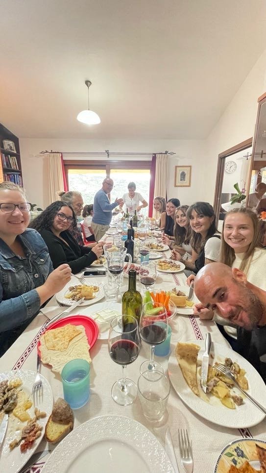 Family and friends gathered around a dining table, enjoying meal and drinks in a well-lit room with large window, curtains, and bookshelf.