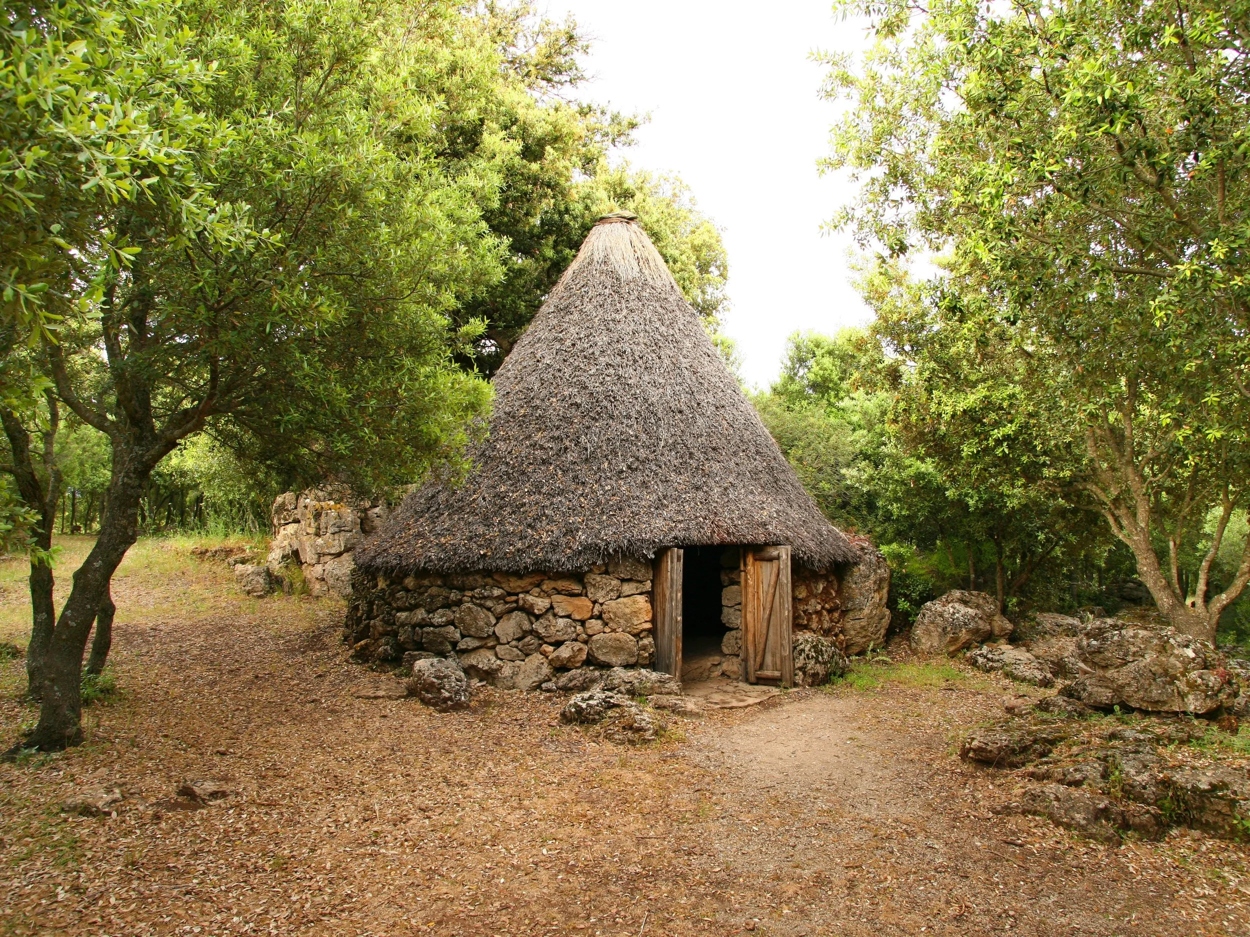 Sardinian traditional round stone and thatch-roof hut surrounded by trees and rocks in a natural setting.