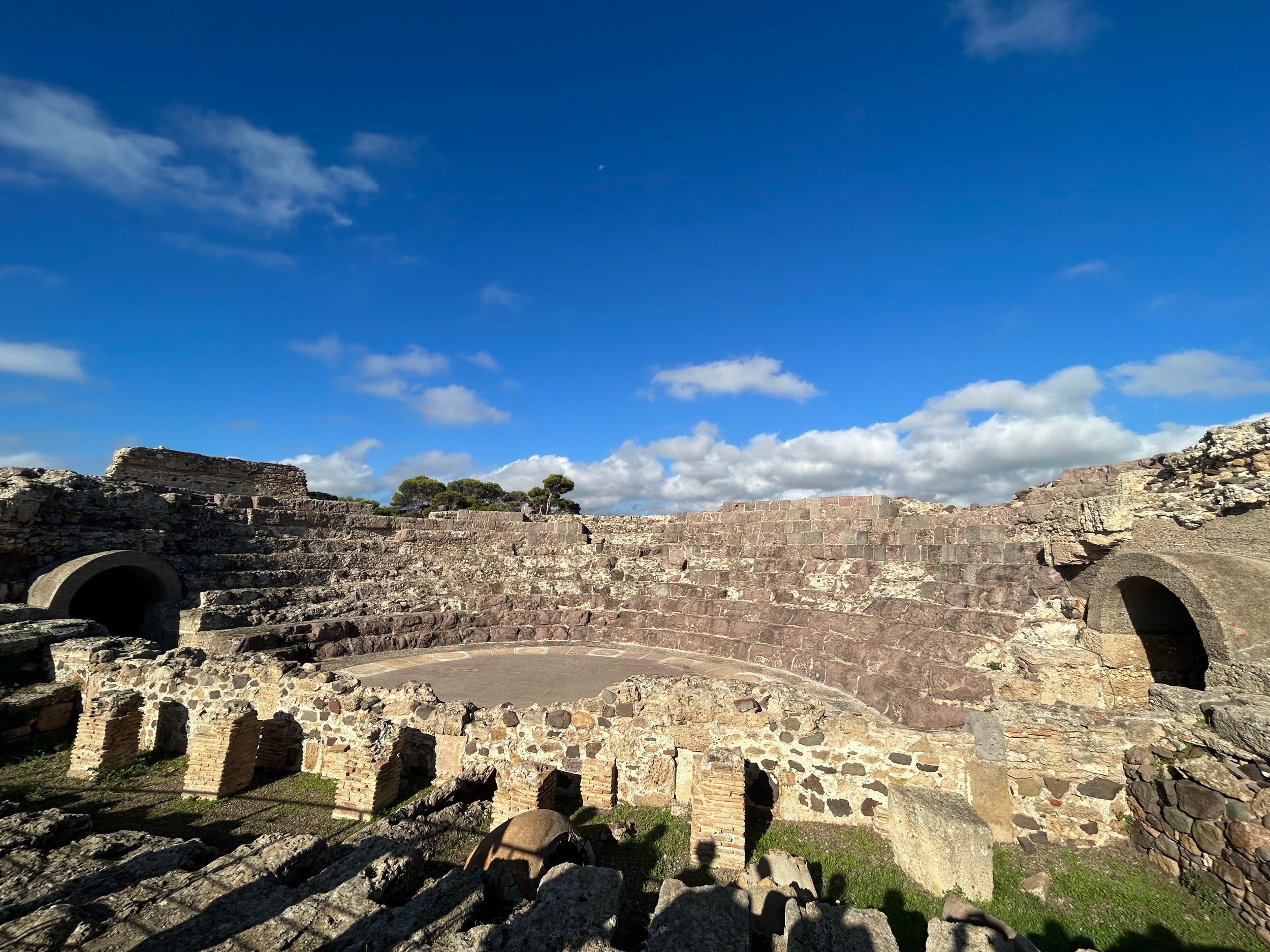 Ancient ruins of a Roman amphitheater made of stone, with arches and steps, under a blue sky with clouds.