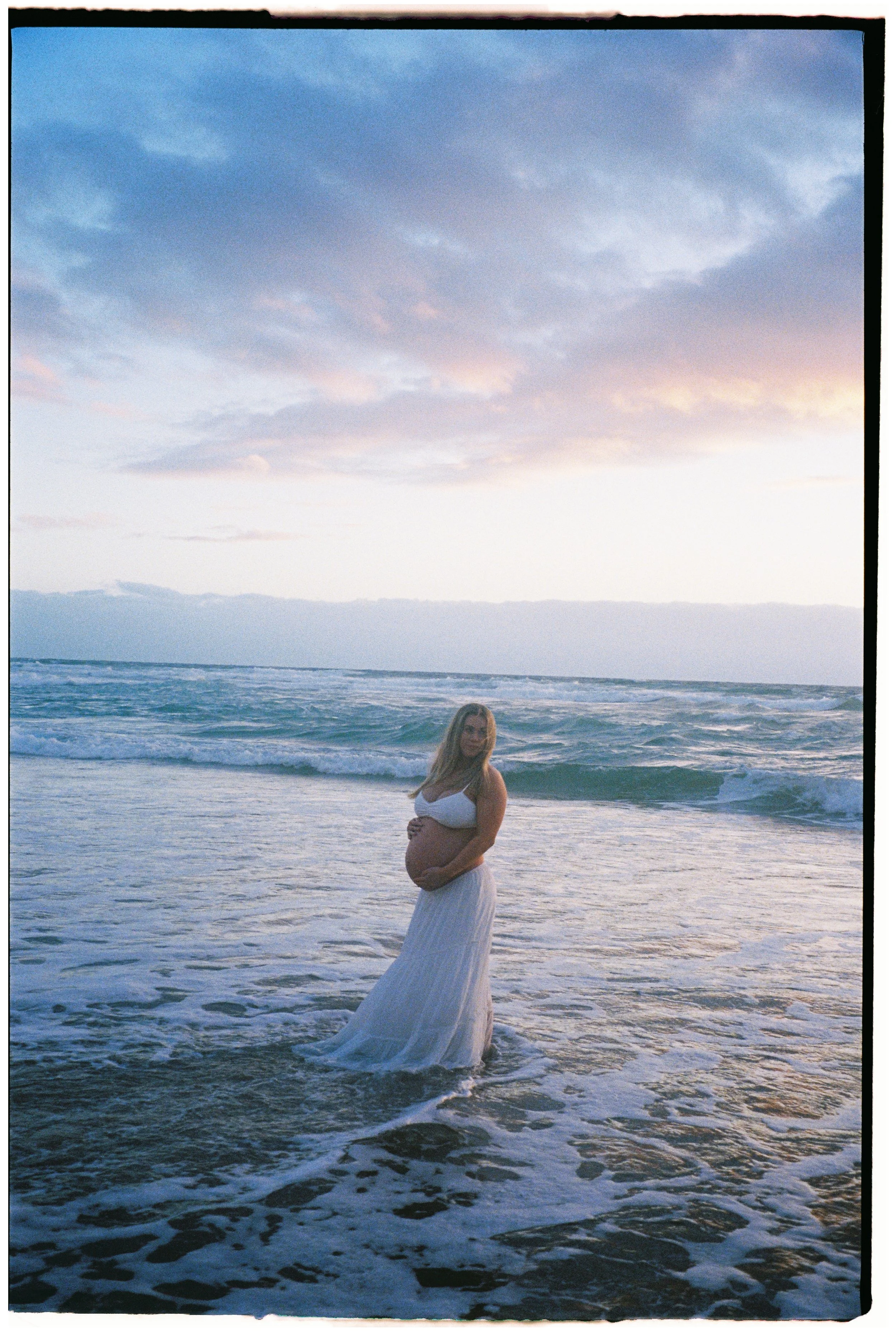 A pregnant woman in a white dress standing in the ocean during sunset or sunrise.