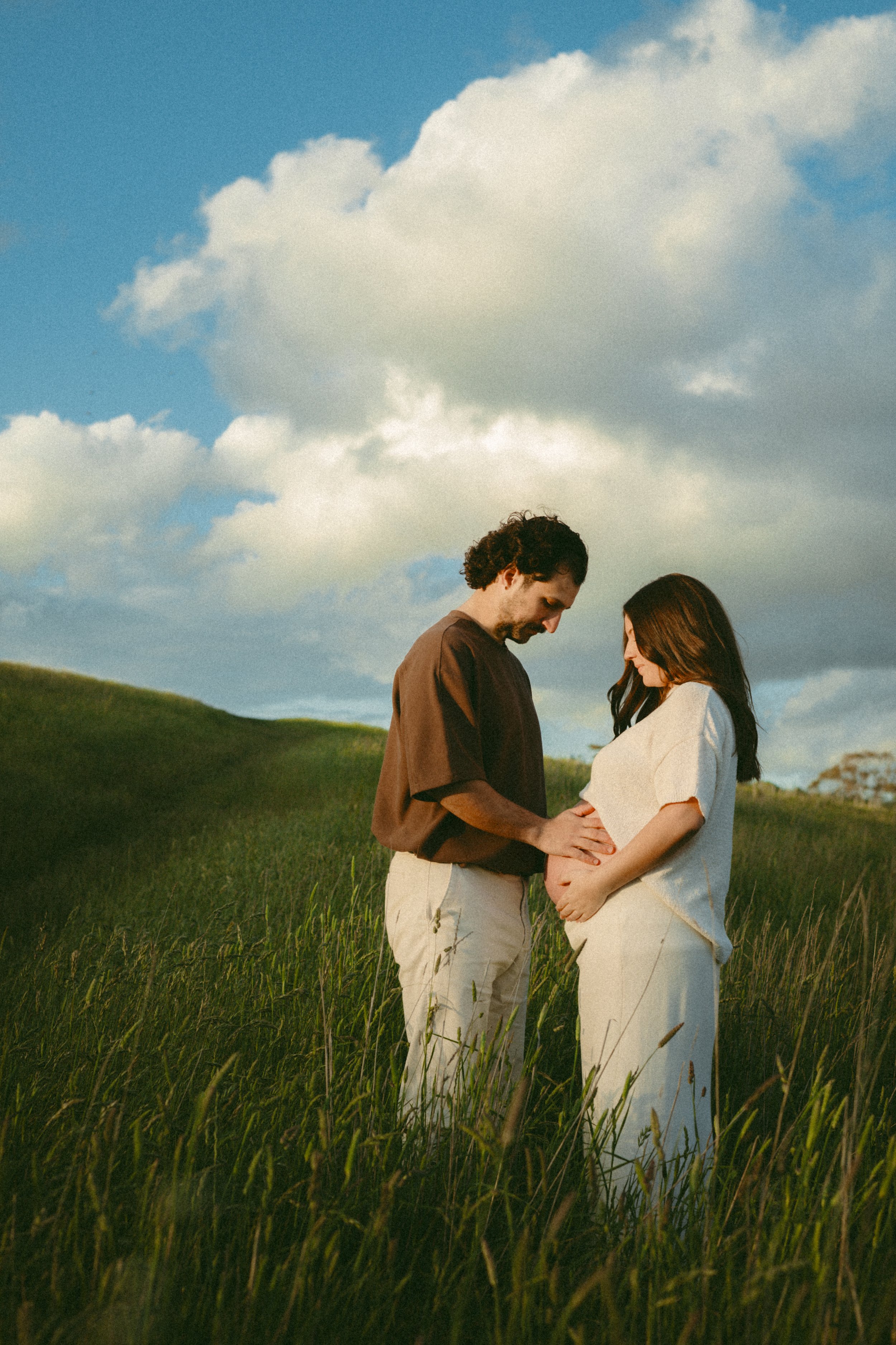A couple standing in a grassy field, with the man touching the woman's pregnant belly, under a partly cloudy sky.