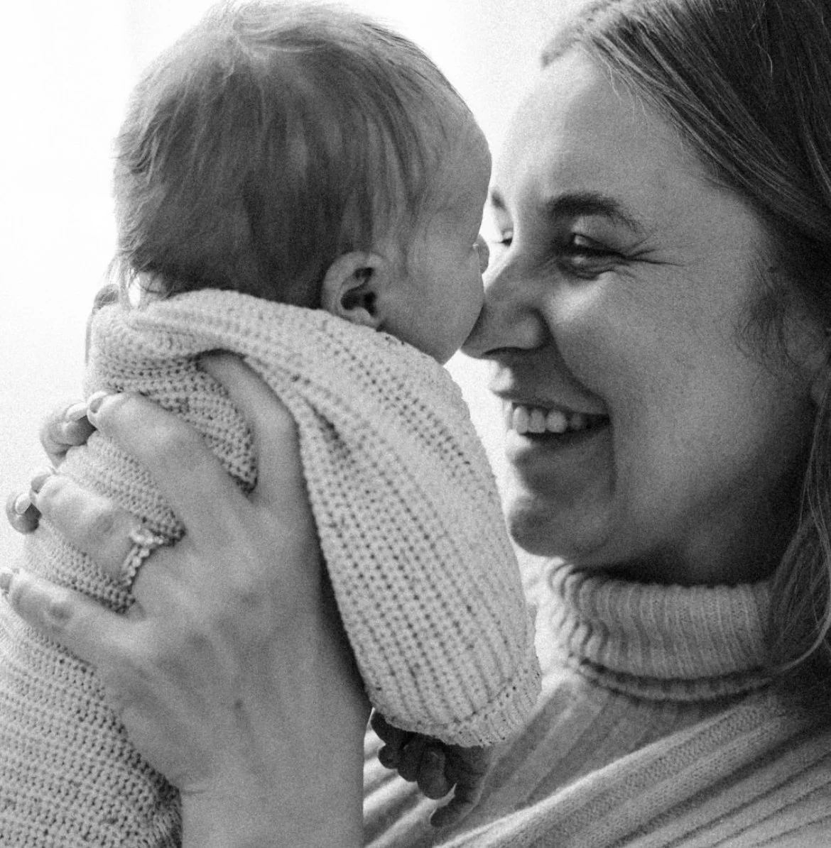 A woman holding a small child close, touching noses, both smiling and making eye contact, in black and white.