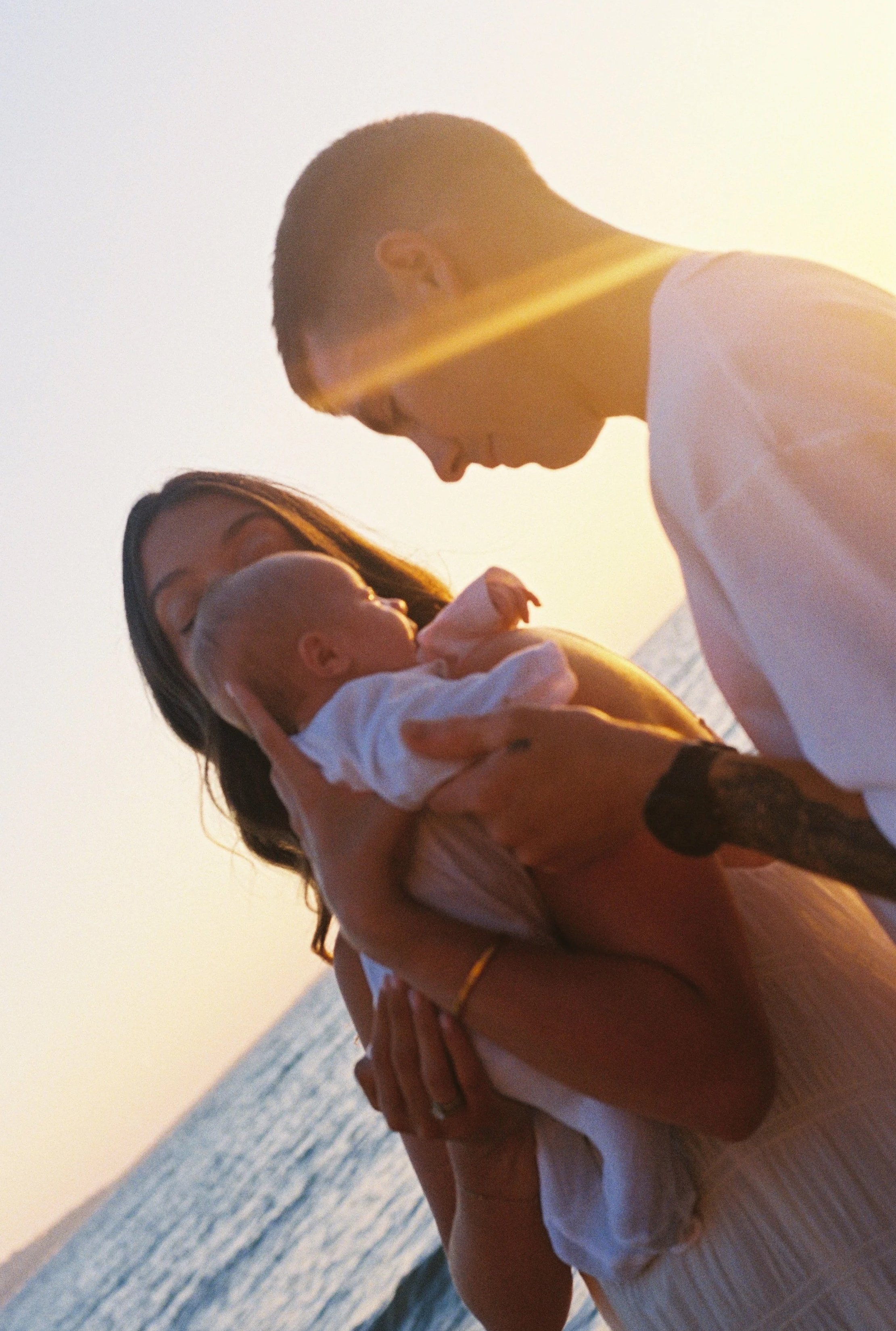 A person holding a baby at the beach during sunset, with the ocean in the background. Maternity, Newborn, Family & Couples Photography | Doreen, Melbourne