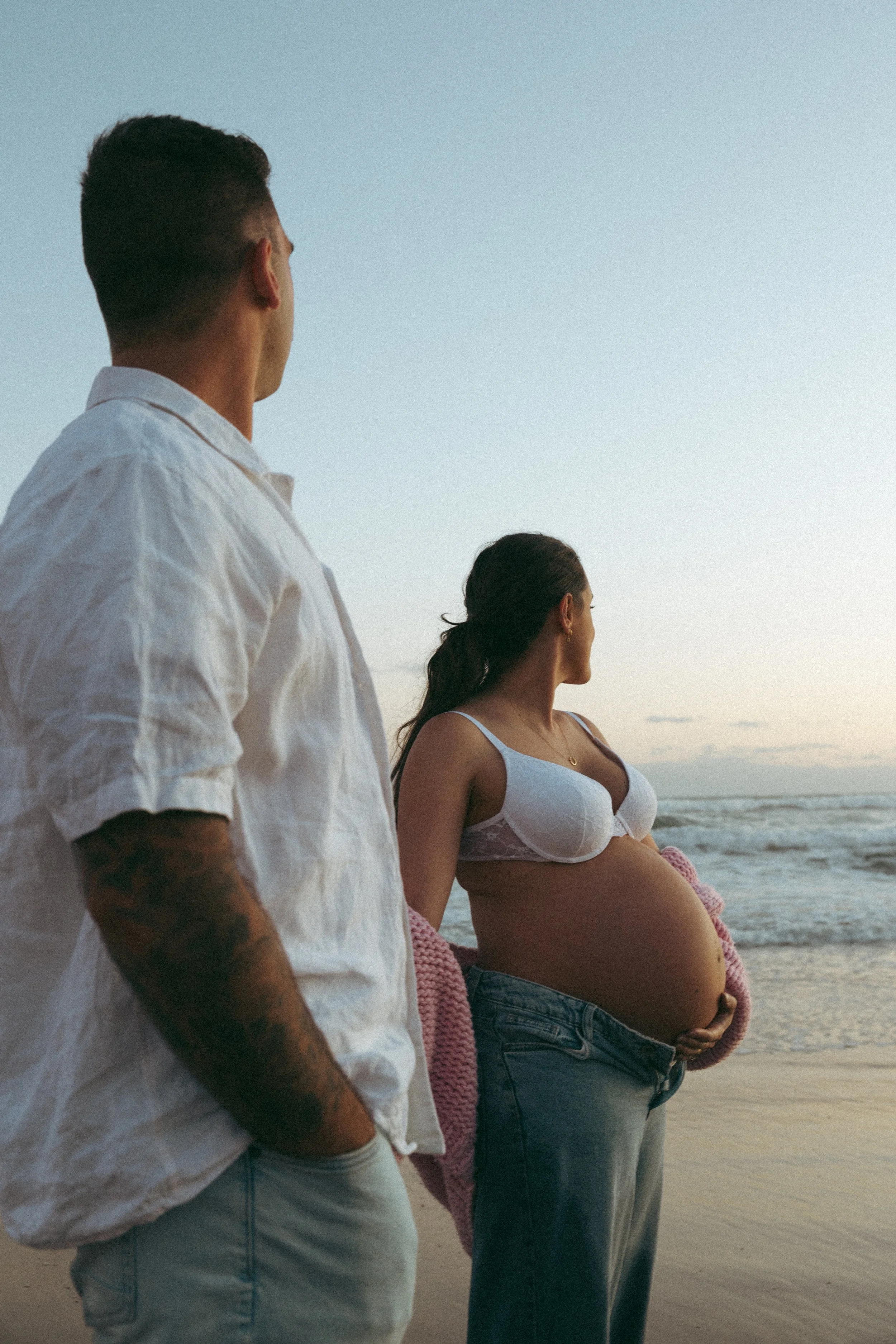 A pregnant woman and a man standing on the beach, looking out at the ocean at sunset.