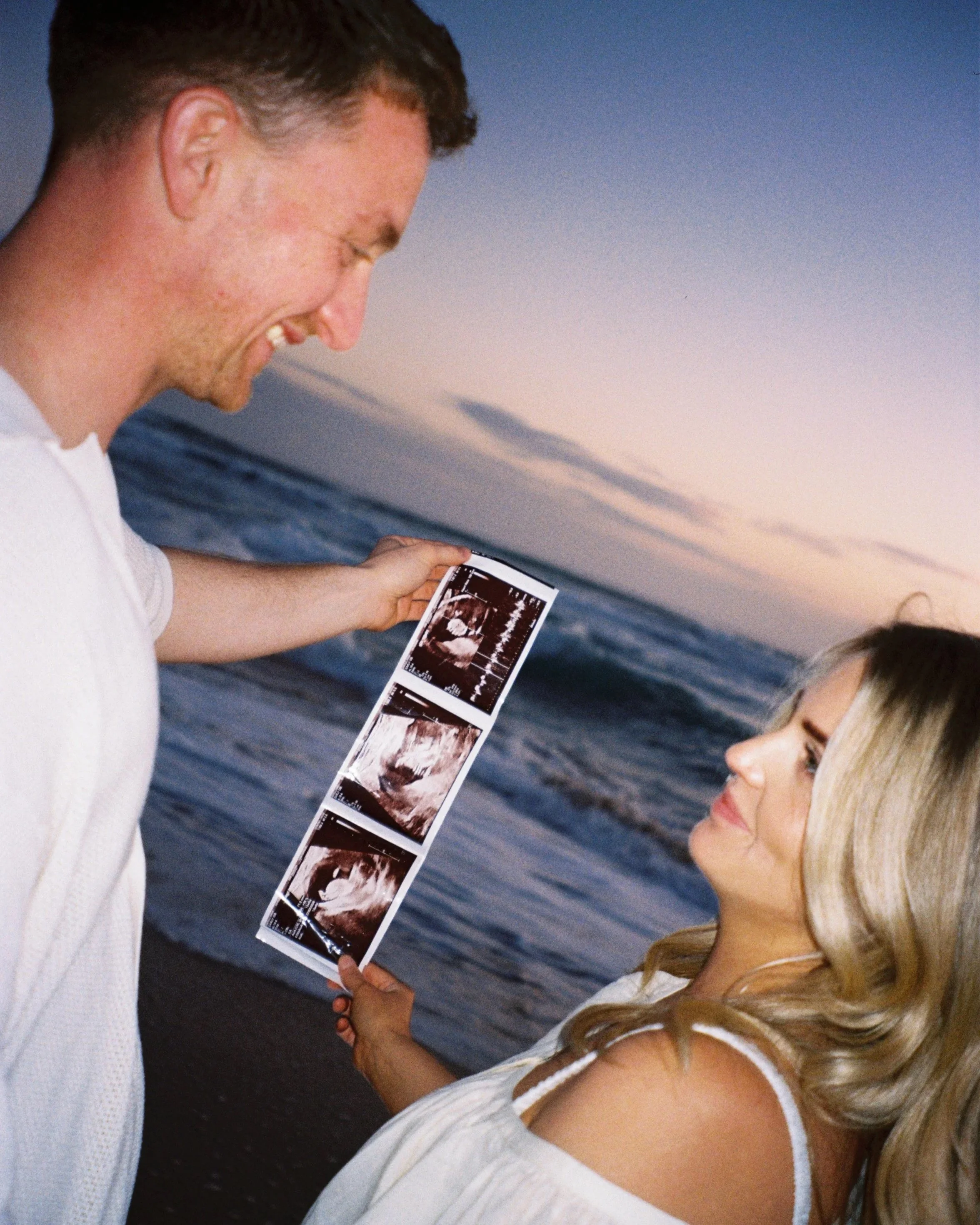 A smiling man is holding ultrasound images in front of a woman on the beach during sunset, with ocean waves in the background.