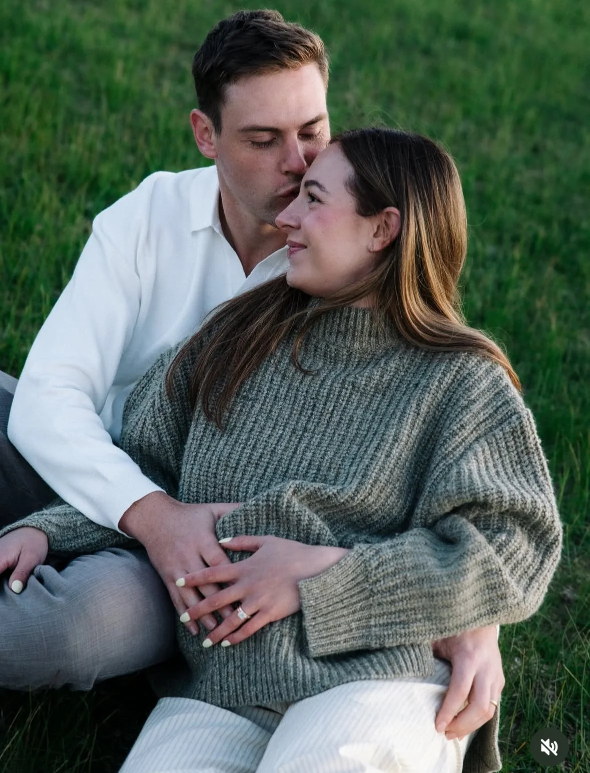 A young man and woman sit close together on grass, with the man gently kissing the woman's forehead and both smiling contentedly.