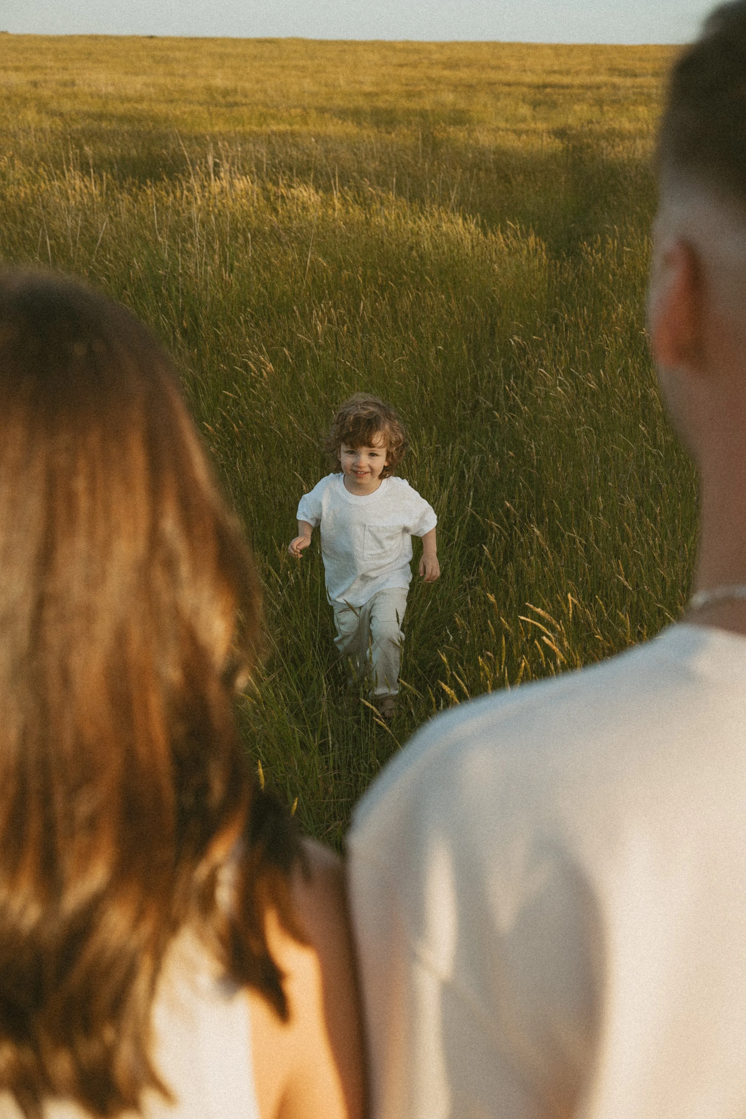 A young boy running through a grassy field during sunset, framed by the blurred backs of two people in the foreground.
Maternity, Newborn, Family & Couples Photography | Doreen, Melbourne