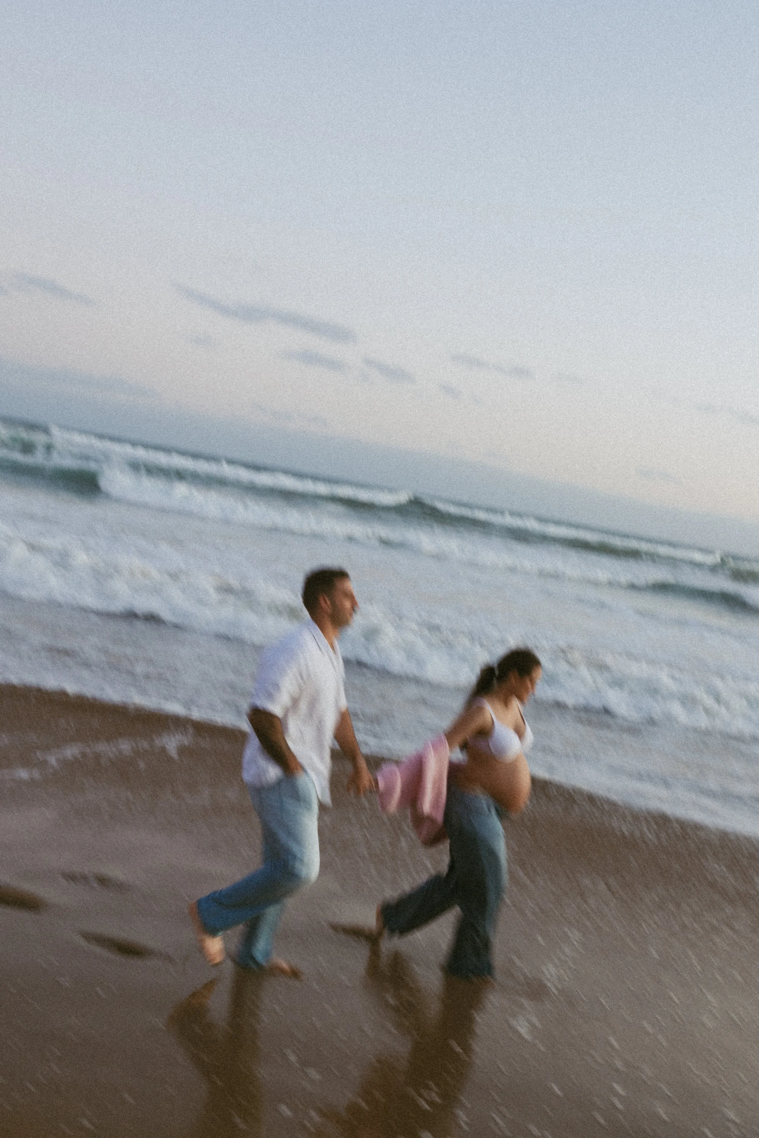 A couple, a pregnant woman, walking hand in hand on the beach during sunset.
