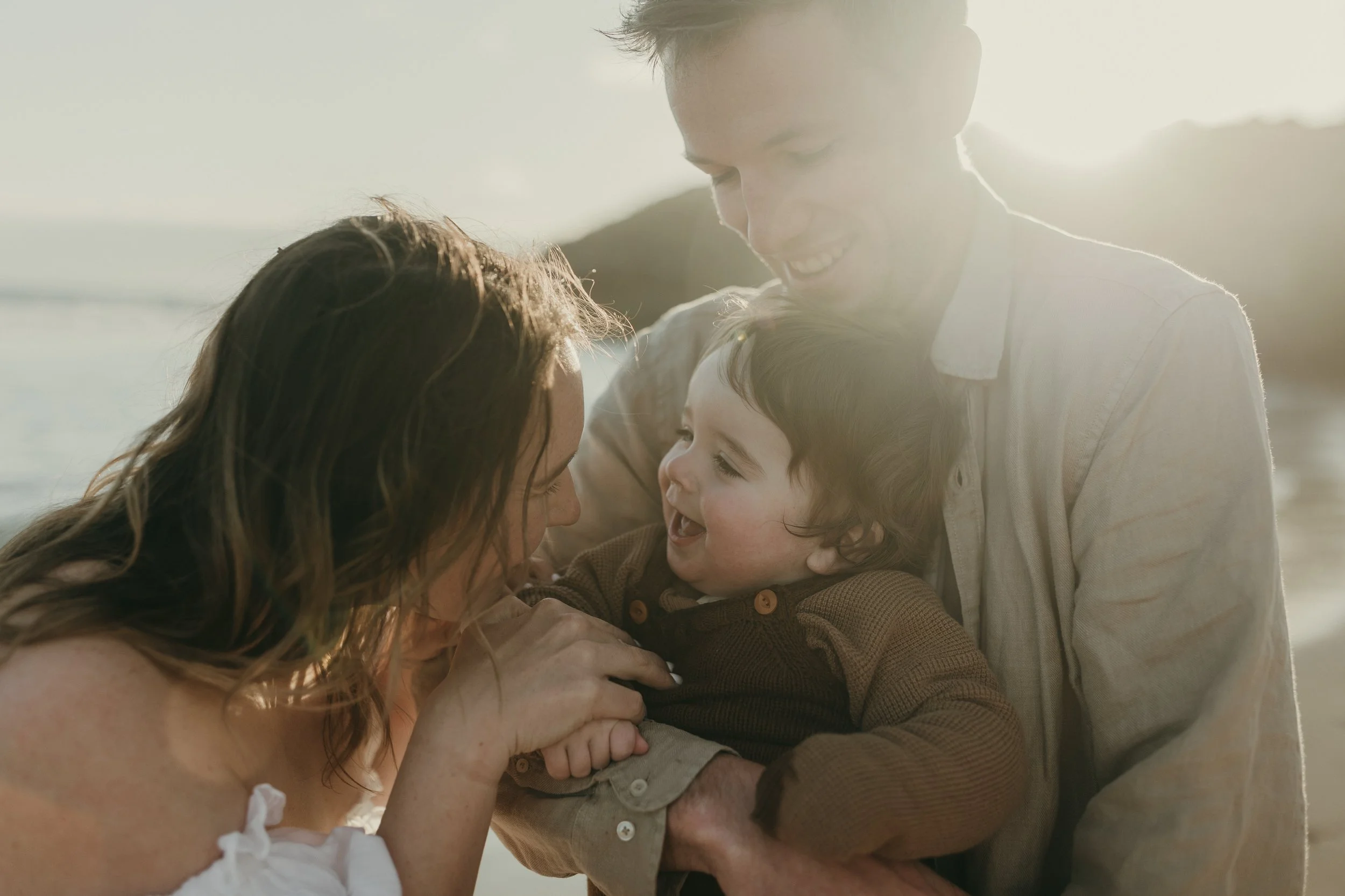 A family of three enjoying a moment together on the beach at sunset. The mother, father, and young boy are smiling and playing closely. The background features a scenic view of water and hills with warm, golden sunlight.