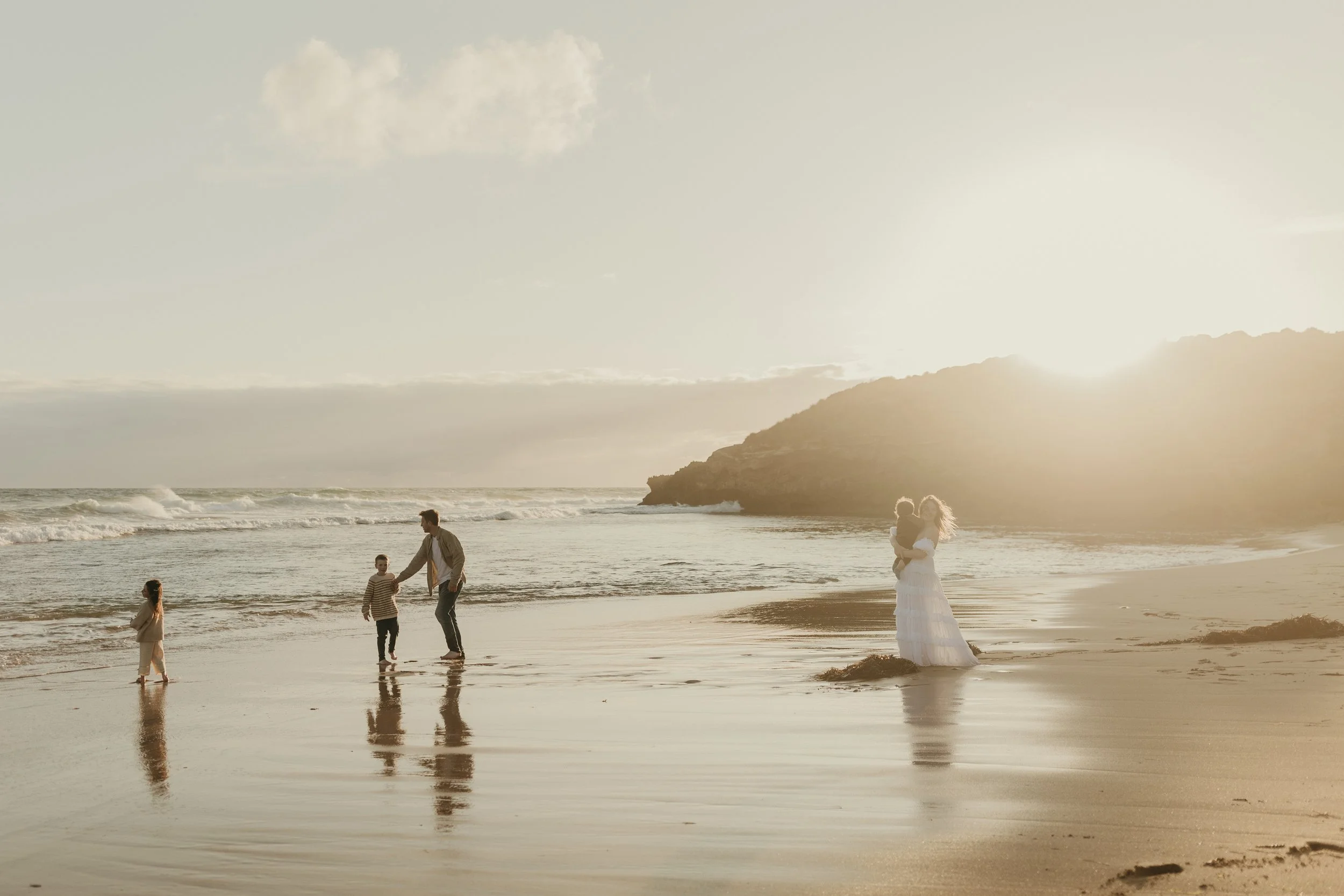 A family on a beach at sunset, with a woman holding a child, two young children playing near the water, and a man with a backpack holding the hand of one child.